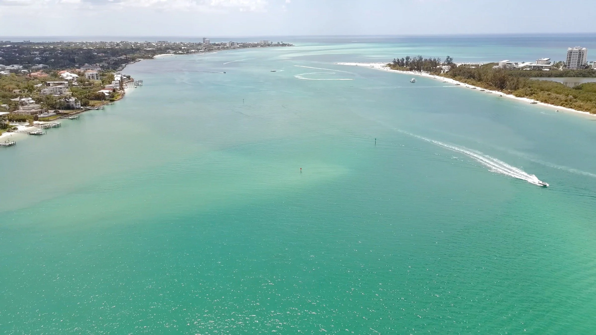 Aerial view of a coastal Sarasota bay with turquoise water, boats, and a shoreline with residential buildings and trees.