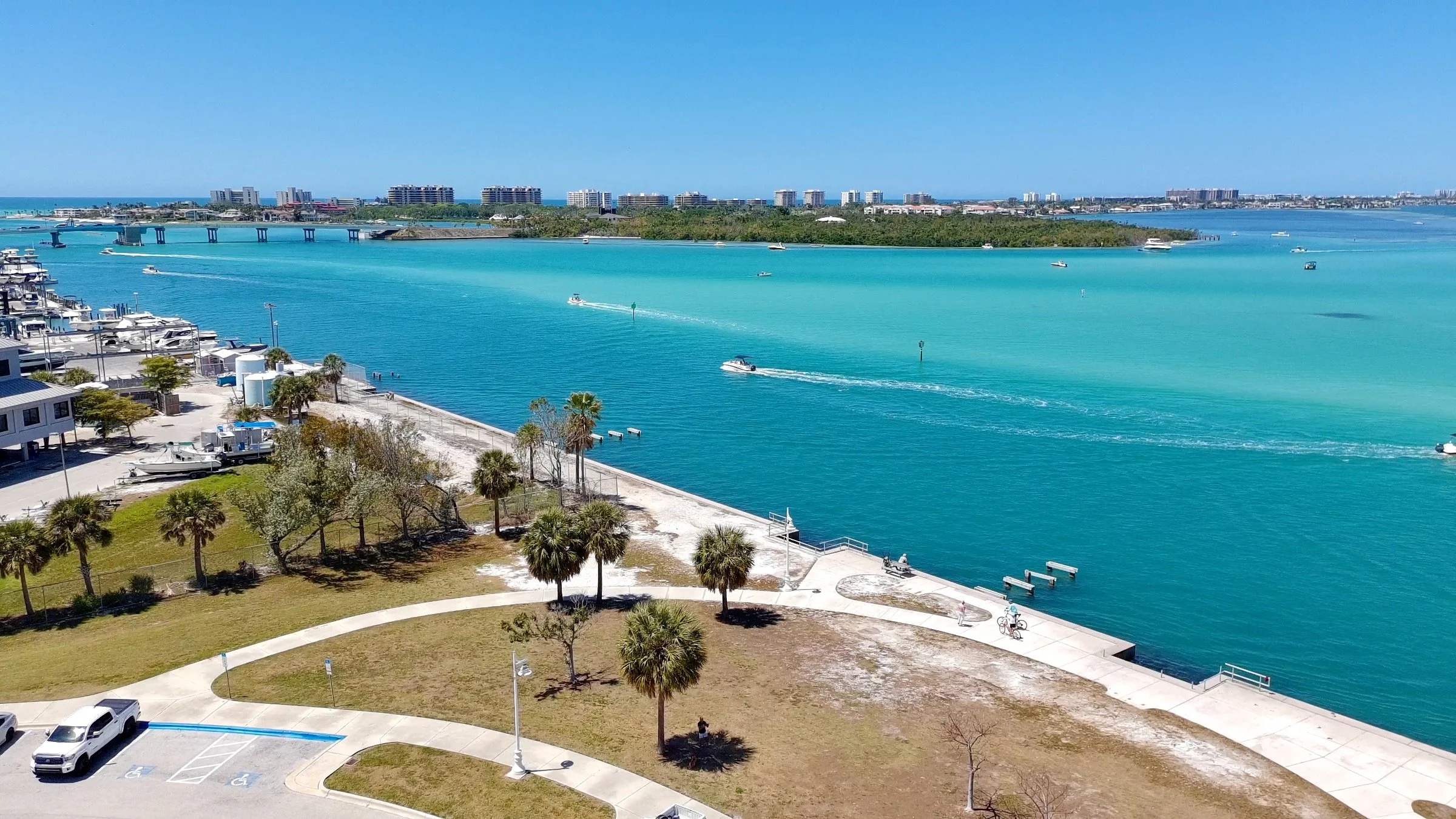 Aerial view of a waterfront park, Sarasota bay, with palm trees, a walking path, parked cars, and boats in the turquoise water, with a city skyline and bridge in the background.