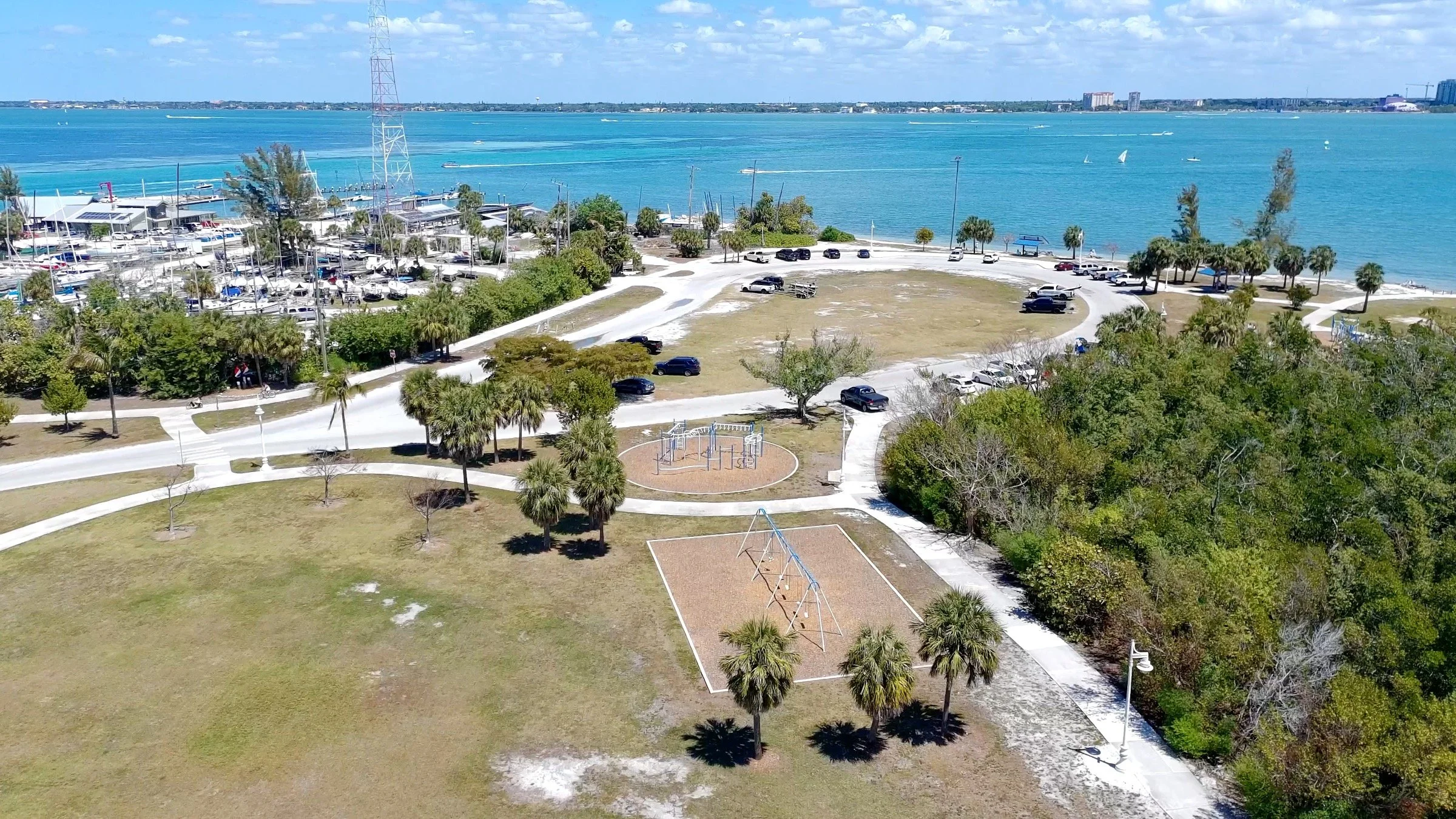 Aerial view of Kensington boat ramps park near a harbor with boats, trees, walking paths, playground, and a parking lot, overlooking a large body of water with sailboats and a city skyline in the background.