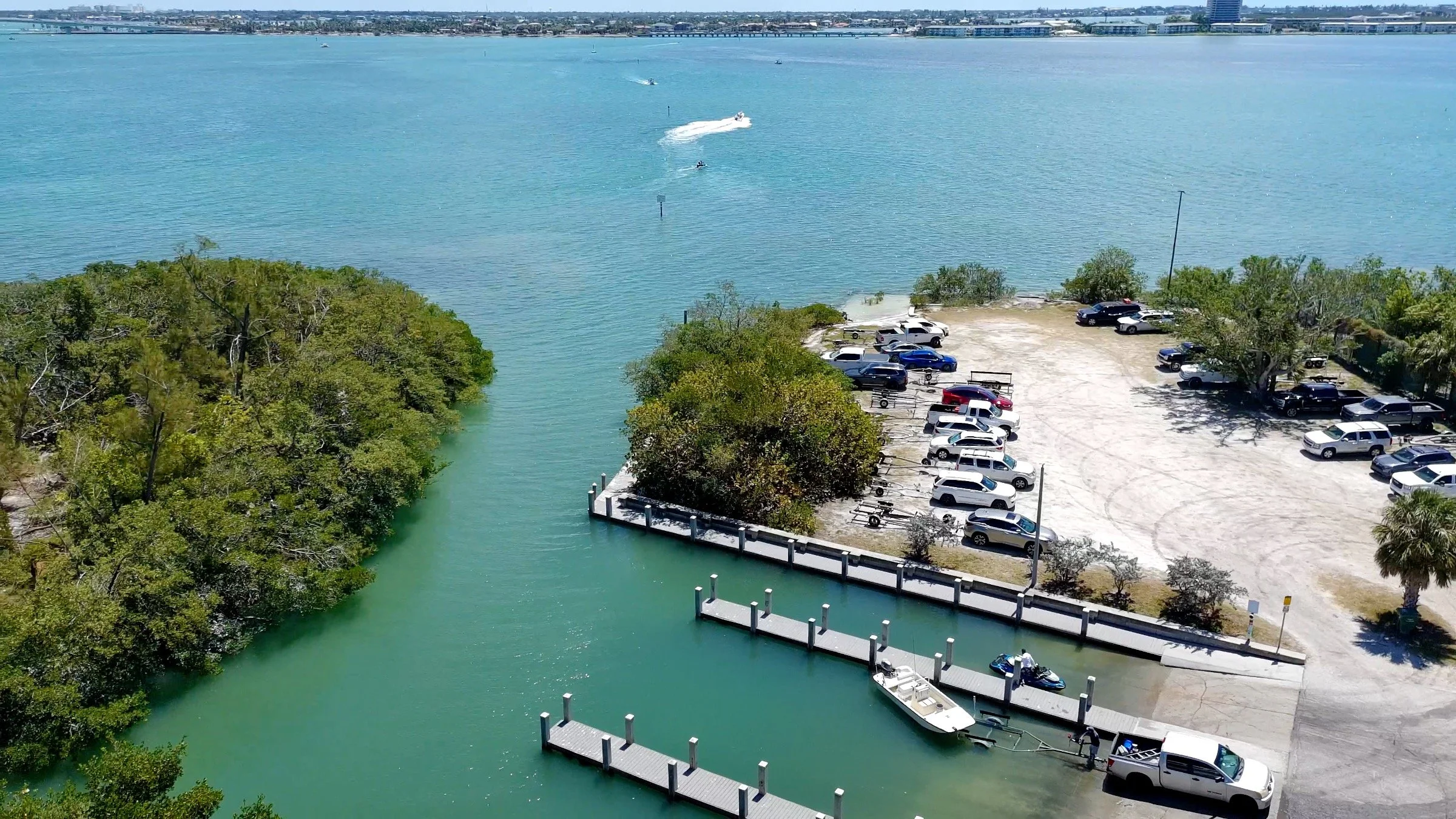 An aerial view of a marina in Sarasota bay with boats docked along piers, adjacent to a parking lot filled with cars, next to a waterway surrounded by green trees, and with a body of water in the background.