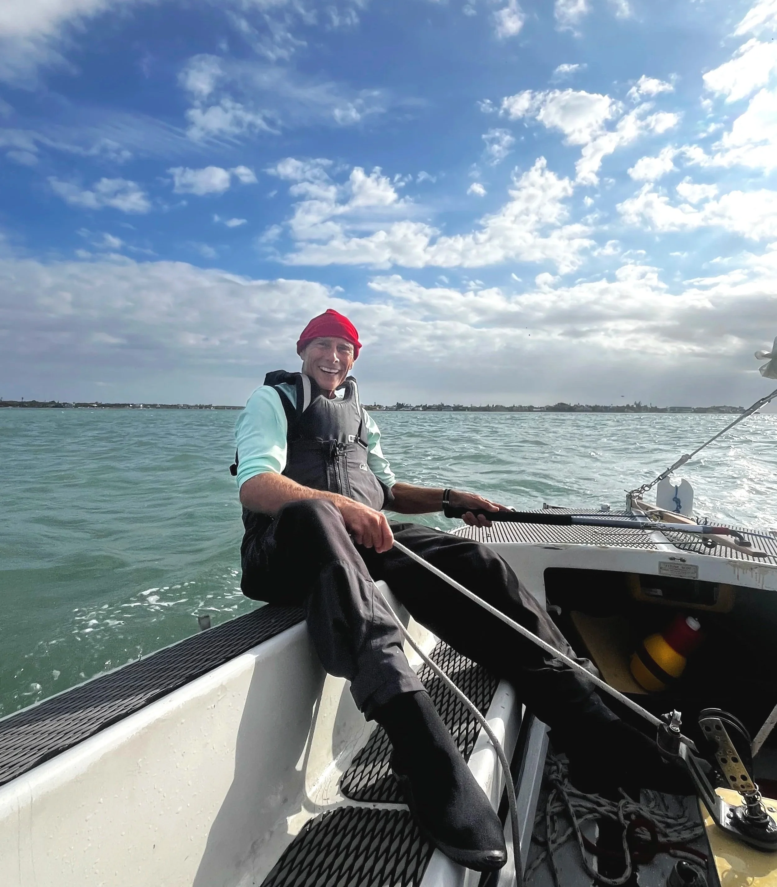 Older man smiling, sitting on the edge of a sailboat, wearing a red beanie, light blue shirt, and black sailing gear, with the ocean and sky in the background.