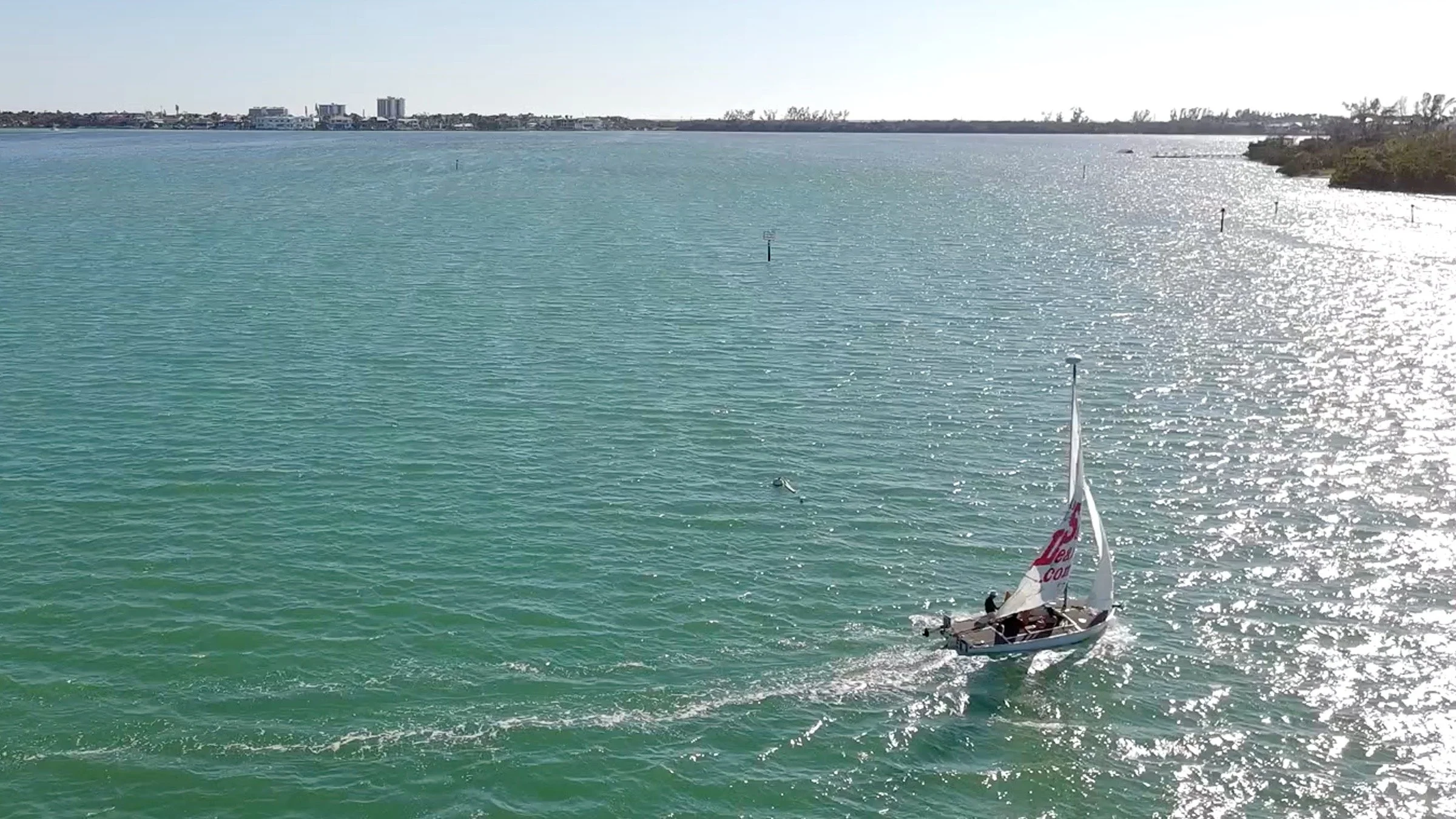 A sailboat on calm, turquoise waters near a shoreline with some trees. In the background, there are buildings across the horizon under a clear sky with sunlight reflecting off the water.