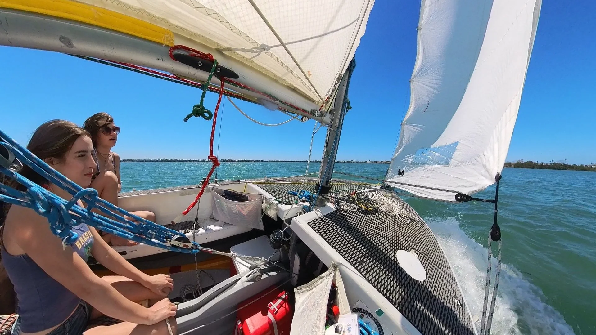 Two women sitting on a small sailboat with sails up, boat moving through water under a clear blue sky.