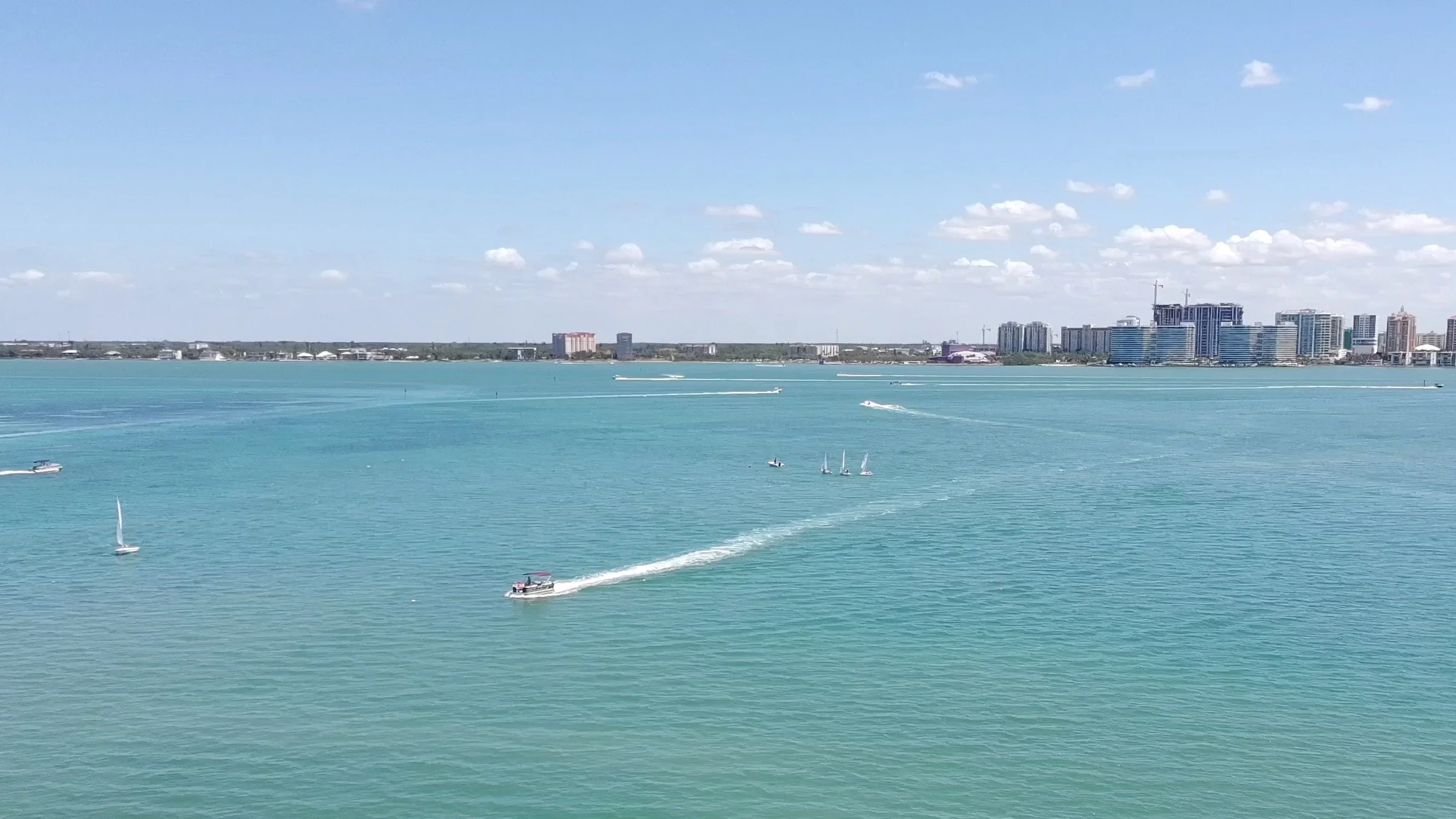 Sarasota Bay city skyline with high-rise buildings across a body of turquoise water with several boats and sailboats, under a partly cloudy sky.