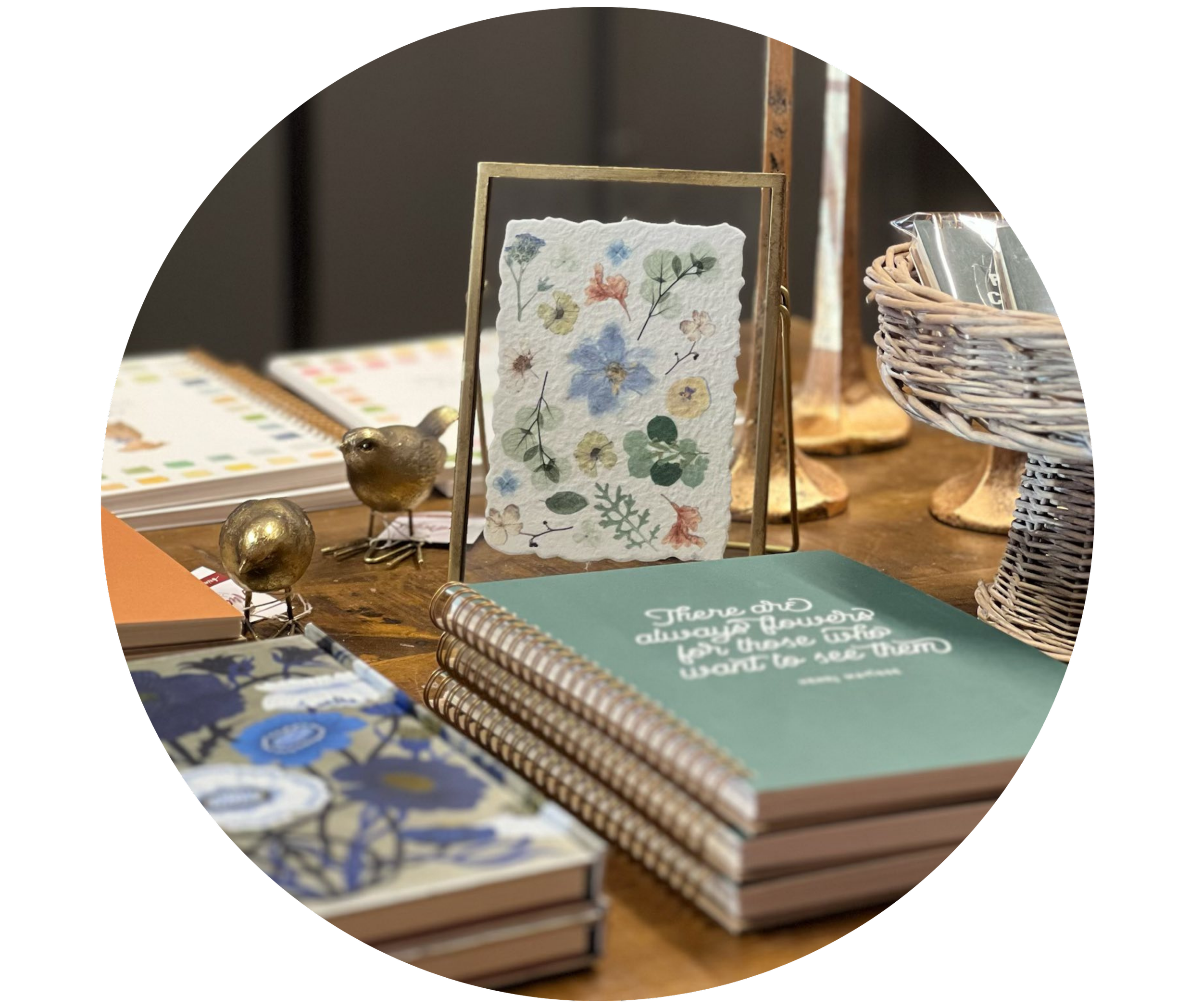 A display of notebooks, a framed botanical illustration on paper, a basket with books, and decorative metallic birds on a wooden table.