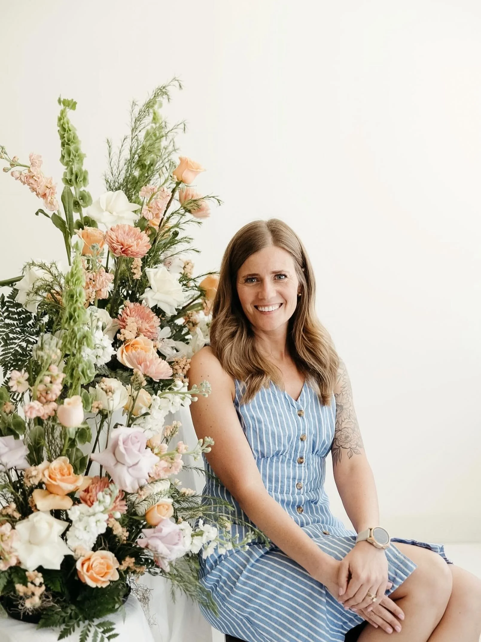 A woman in a blue striped dress sitting next to a large bouquet of mixed flowers against a plain white background.