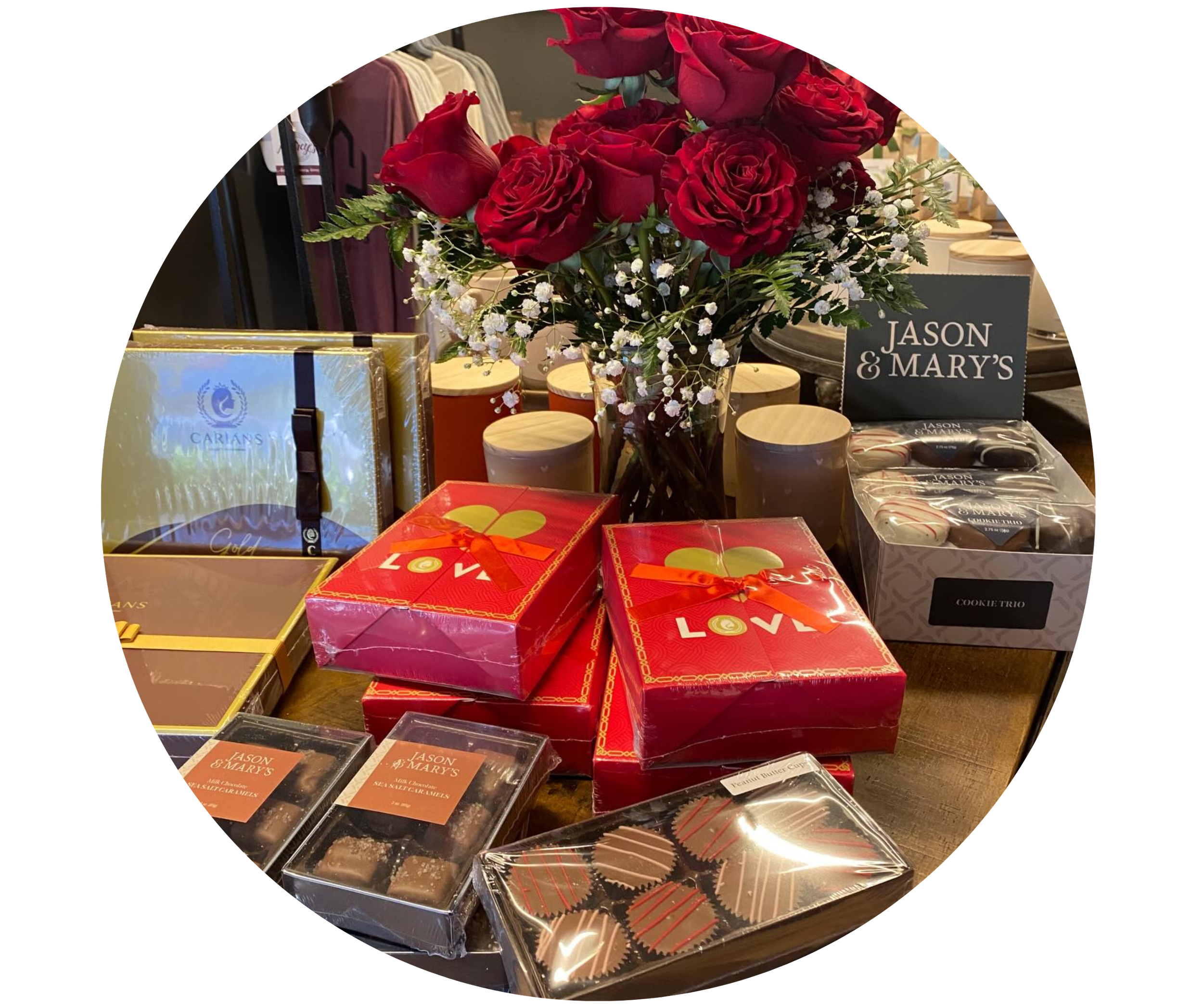 A floral arrangement of red roses, lilies, and white baby's breath in a clear glass vase, surrounded by boxes of chocolates and gifts on a wooden table.