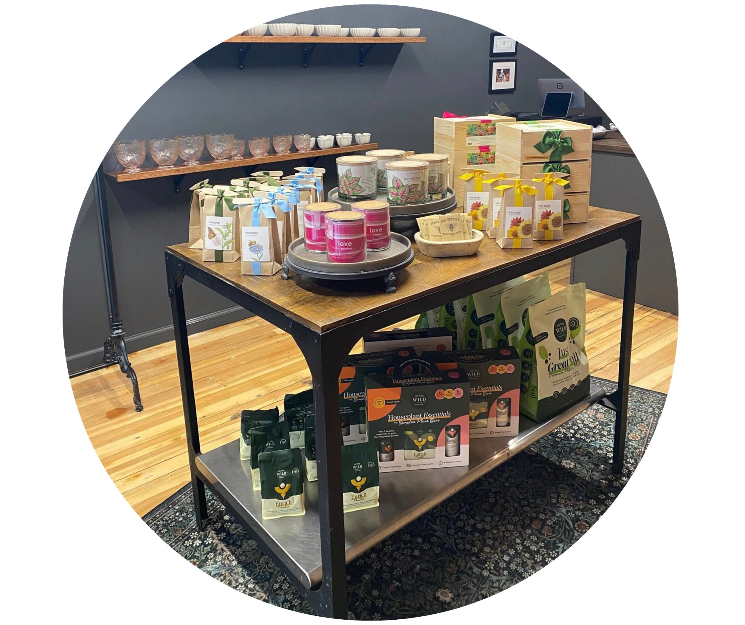 Display table with various packaged products including food items, candles, and plant-based products in a retail store, with wall shelves and framed pictures in the background.