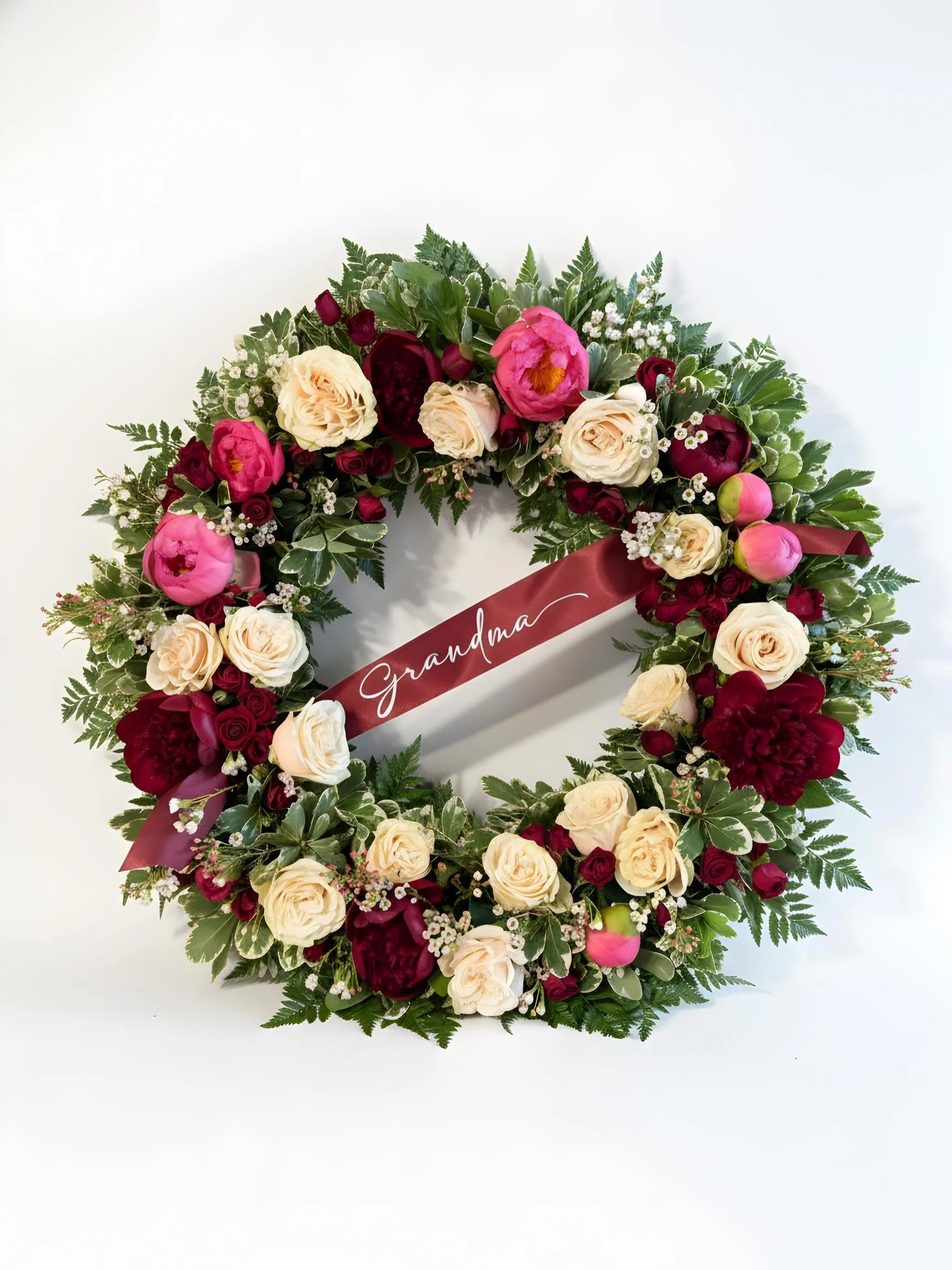 A floral wreath with pink, cream, and deep red flowers and greenery, featuring a maroon ribbon with the word 'Grandma' written on it.