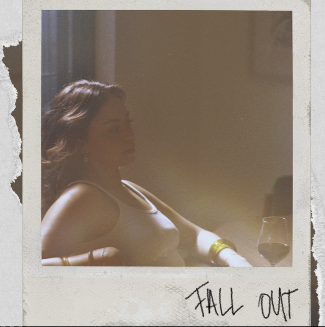 A woman with wavy hair sitting at a table with a glass of red wine, her eyes closed or looking down, with sunlight casting a warm glow on her face. The photo has a Polaroid-style border with the words "FALL OUT" handwritten at the bottom.