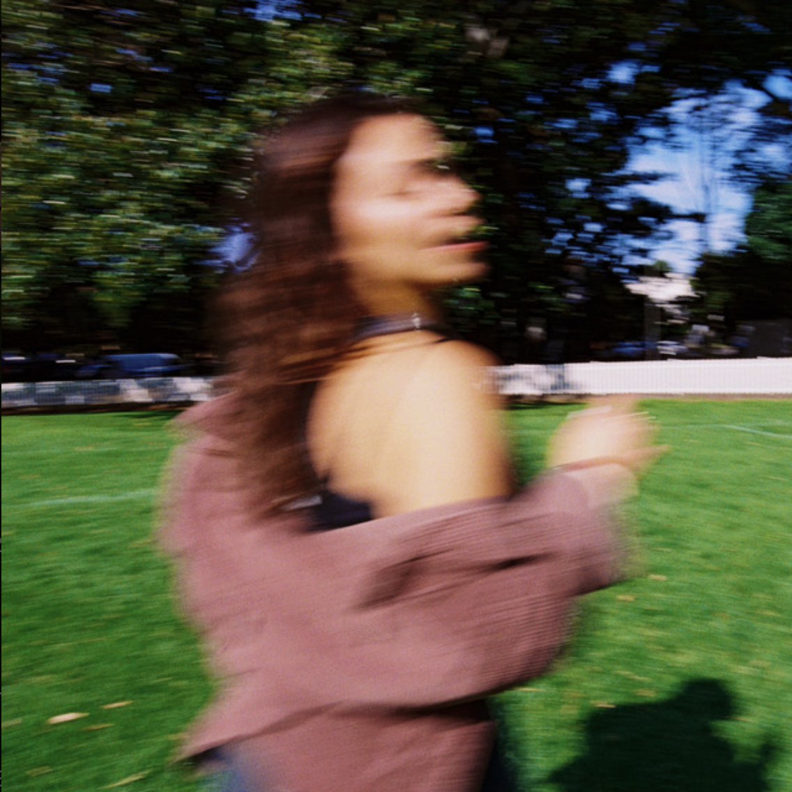 Blurred image of a woman with long brown hair running outdoors on a grassy field with trees and blue sky in the background.