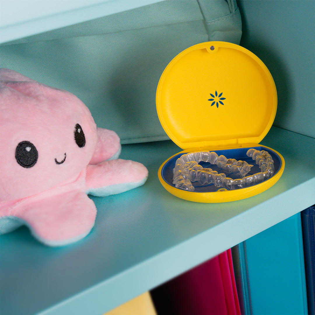 Pink plush octopus toy with smiling face and big black eyes next to a yellow case with a blue interior, holding a clear dental retainer or mouthguard on a light blue shelf with colorful folders underneath.