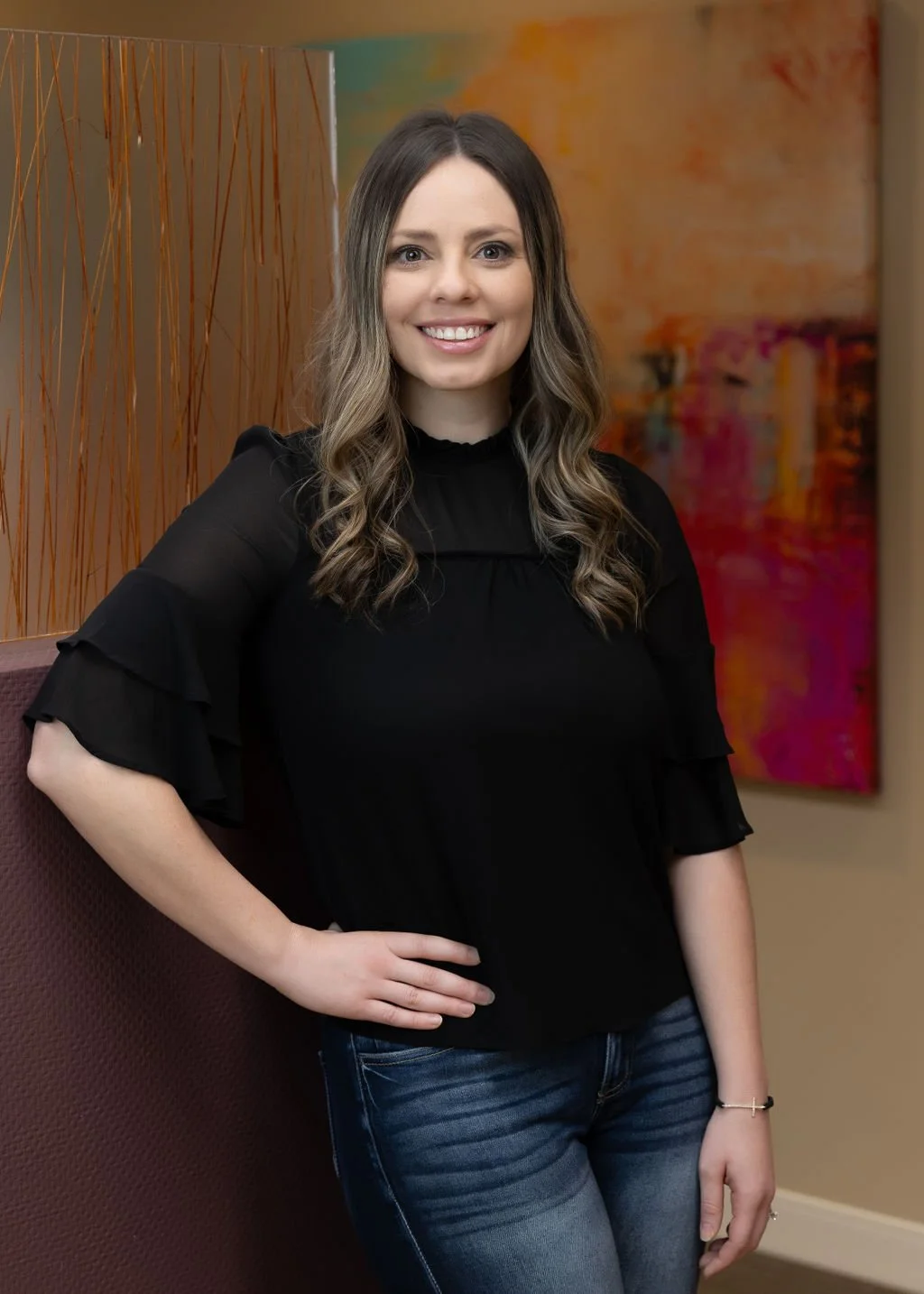 A smiling woman with long wavy brown hair wearing a black top and blue jeans, standing in front of colorful abstract art and a decorative wooden partition.