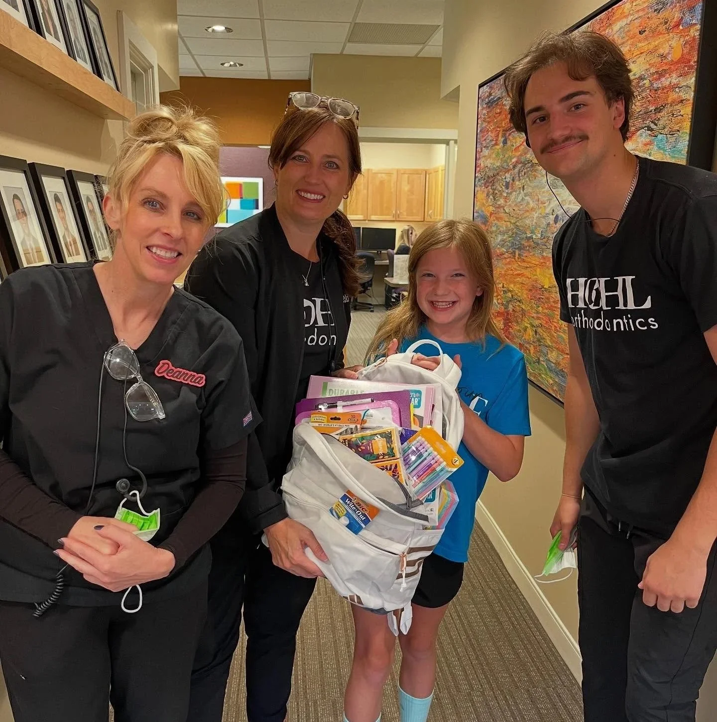 A young girl holding a backpack filled with school supplies, smiling with three adults, in an indoor setting with framed pictures and artwork on the walls.