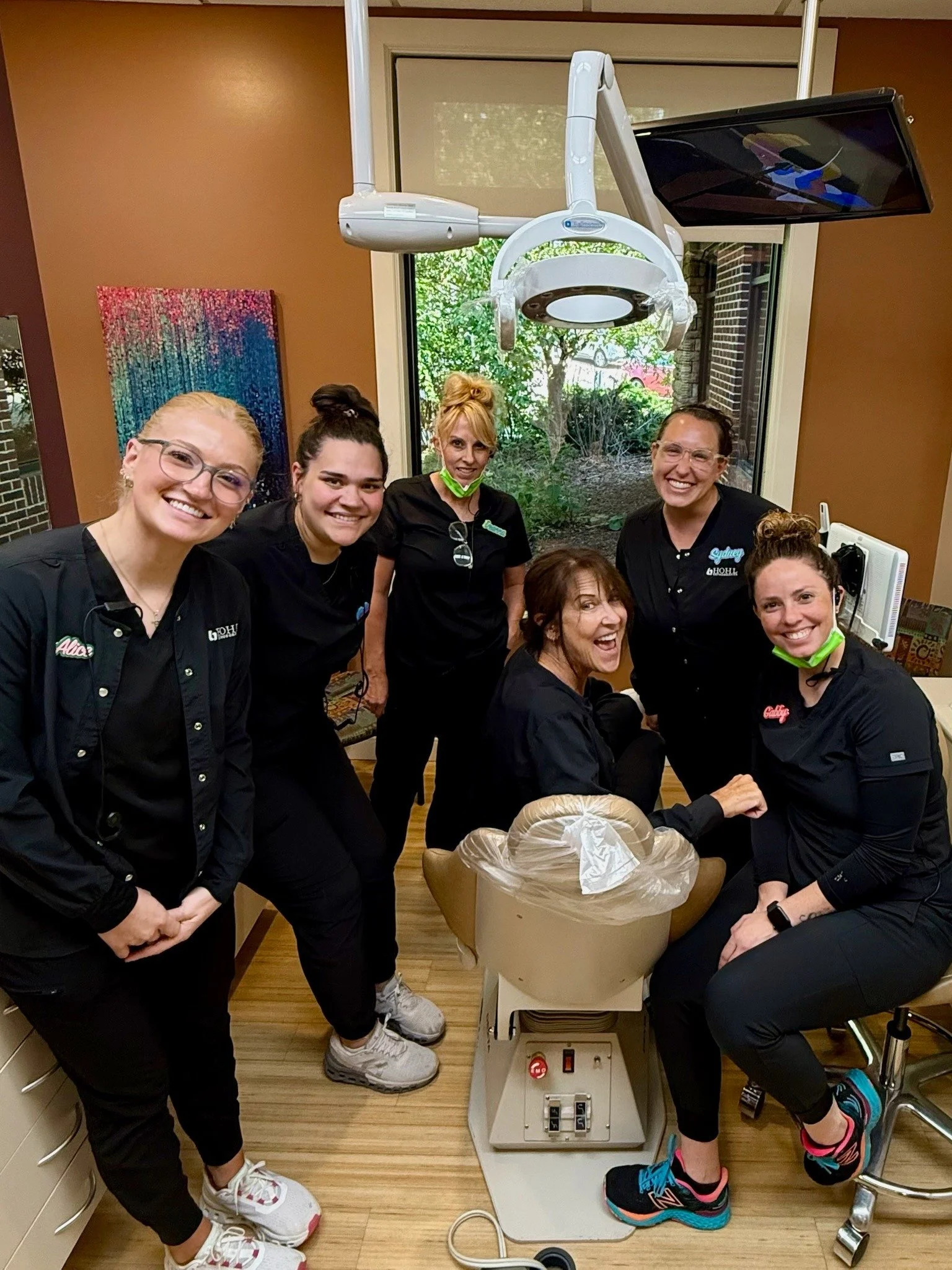 Group of six women in black uniforms gathered around a dental chair inside a dental office. The woman sitting in the chair is smiling and the others are standing or sitting nearby, smiling. There is dental equipment and a window with greenery outside.