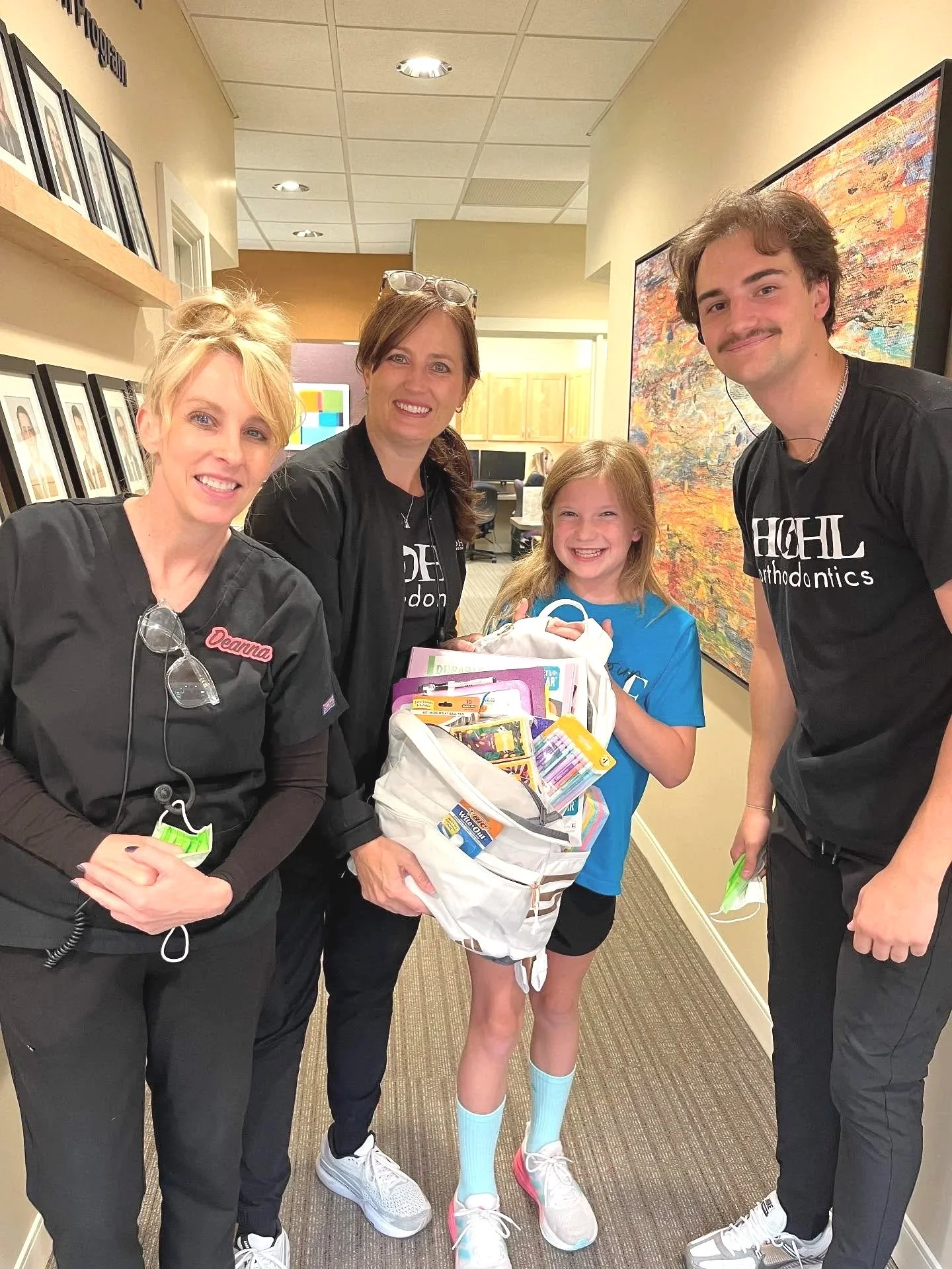 A young girl holding a bag full of school supplies and smiling, standing with three adults in a hallway of a clinic or office building. The adults are wearing black scrubs with the word 'Highland Orthodontics' and a name tag, and there are framed pictures on the wall behind them.