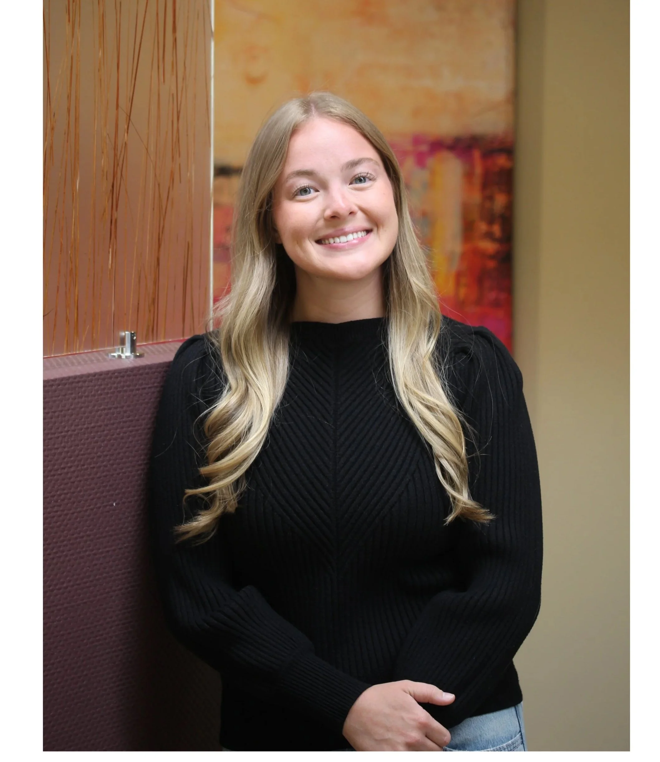A young woman with long blonde hair, smiling and wearing a black sweater, standing indoors with a colorful abstract painting in the background and a maroon chair in the foreground.