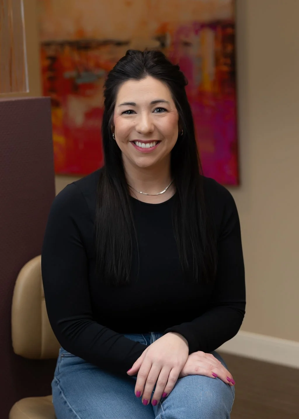A smiling woman with long dark hair, wearing a black top and blue jeans, sitting in front of an abstract colorful painting.