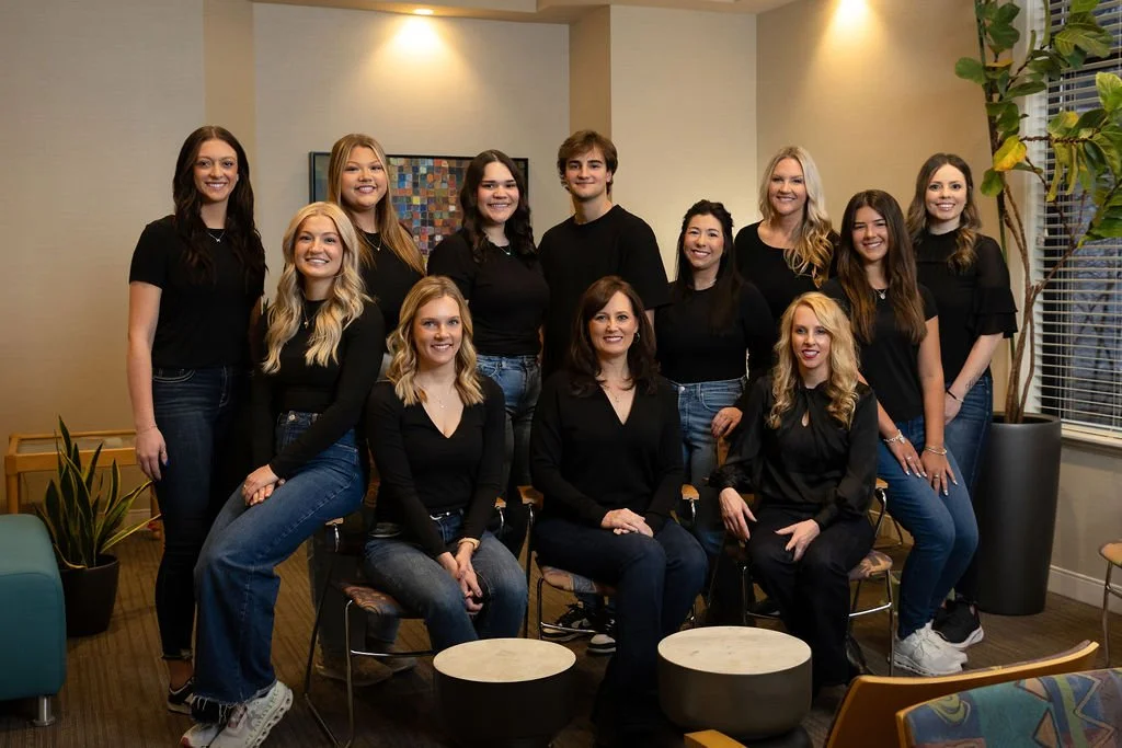 Group of thirteen women and one man smiling in a modern office space with plants and artwork.