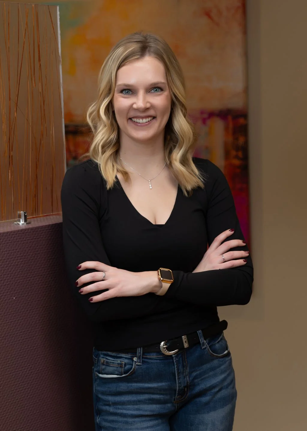 A young woman with blonde hair wearing a black top and blue jeans, standing with arms crossed, smiling in an indoor setting with colorful abstract artwork in the background.