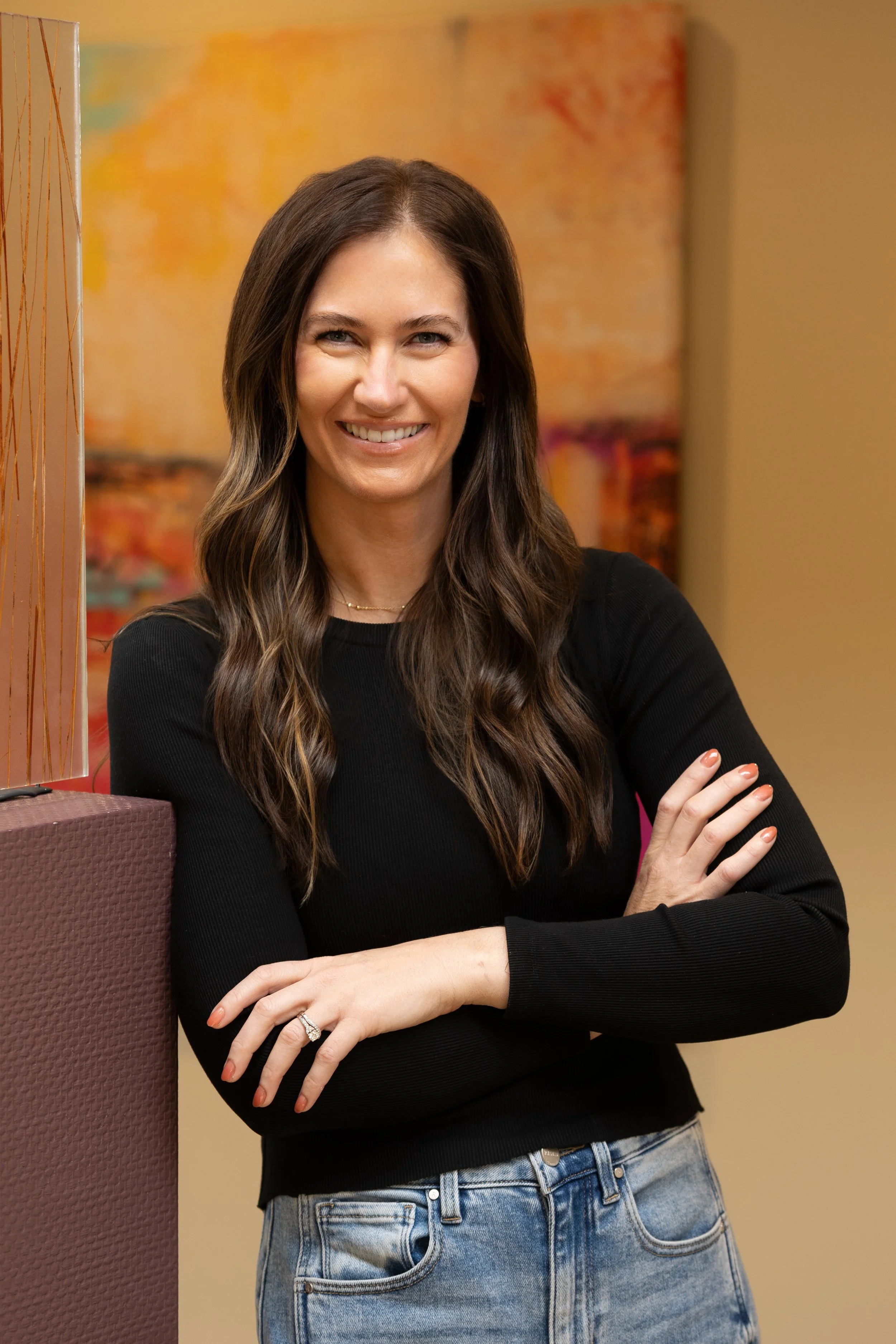 A woman with long, wavy brown hair smiling and standing with crossed arms, wearing a black long-sleeve top and blue jeans, in an indoor setting with colorful abstract art in the background.