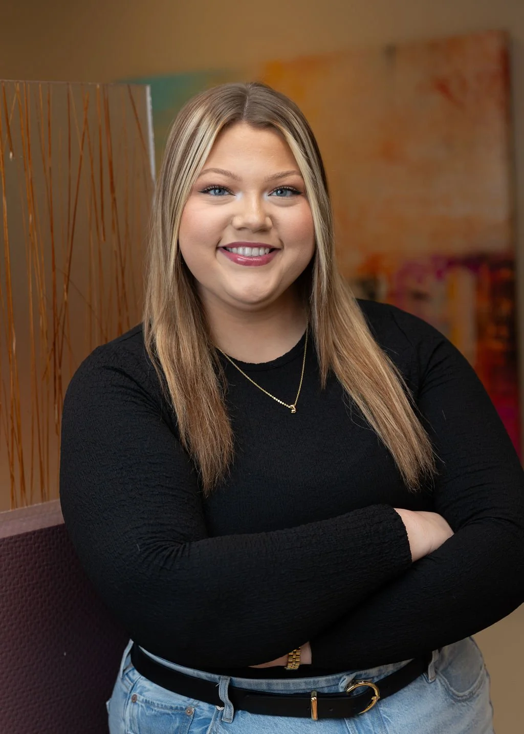 A young woman with blonde hair and blue eyes smiling, standing with arms crossed in front of a decorative room divider with an abstract colorful background.