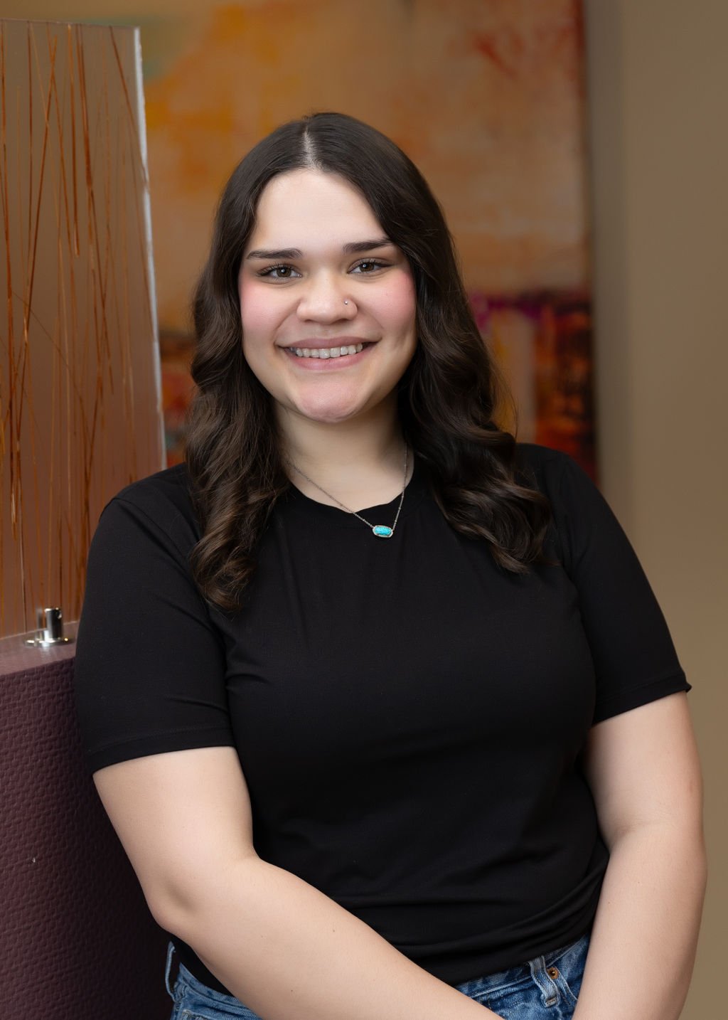 Young woman with long dark hair, smiling, wearing a black shirt and jewelry, sitting indoors.