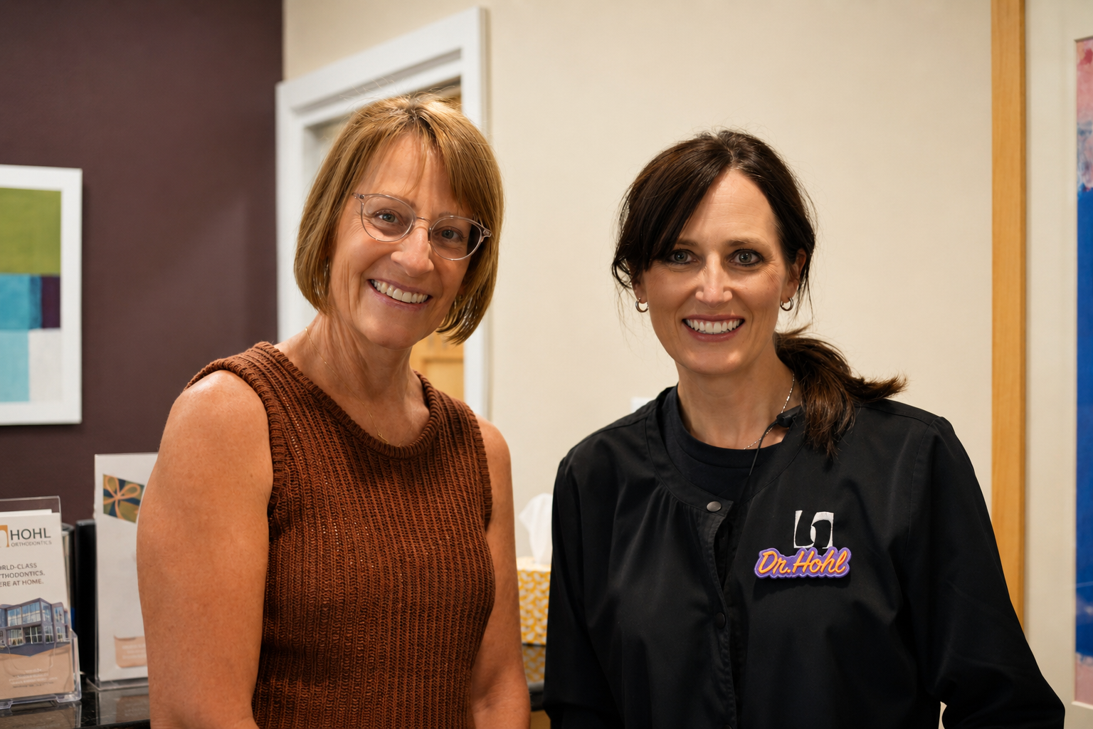 Two women smiling in an indoor setting, one with short red hair and glasses wearing a sleeveless brown top, and the other with long dark hair wearing a black jacket with a name tag that says 'Dr. Hohl'.