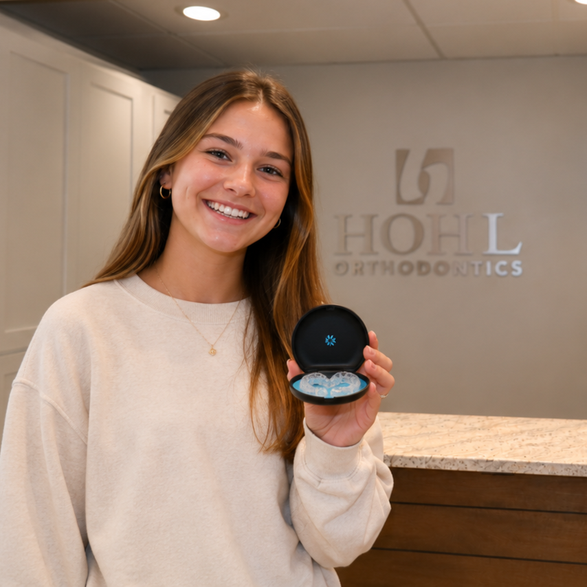 A young woman with long, light brown hair smiling and holding a small black case with blue dental aligners inside, standing in a dental office with the sign 'HOHL Orthodontics' in the background.