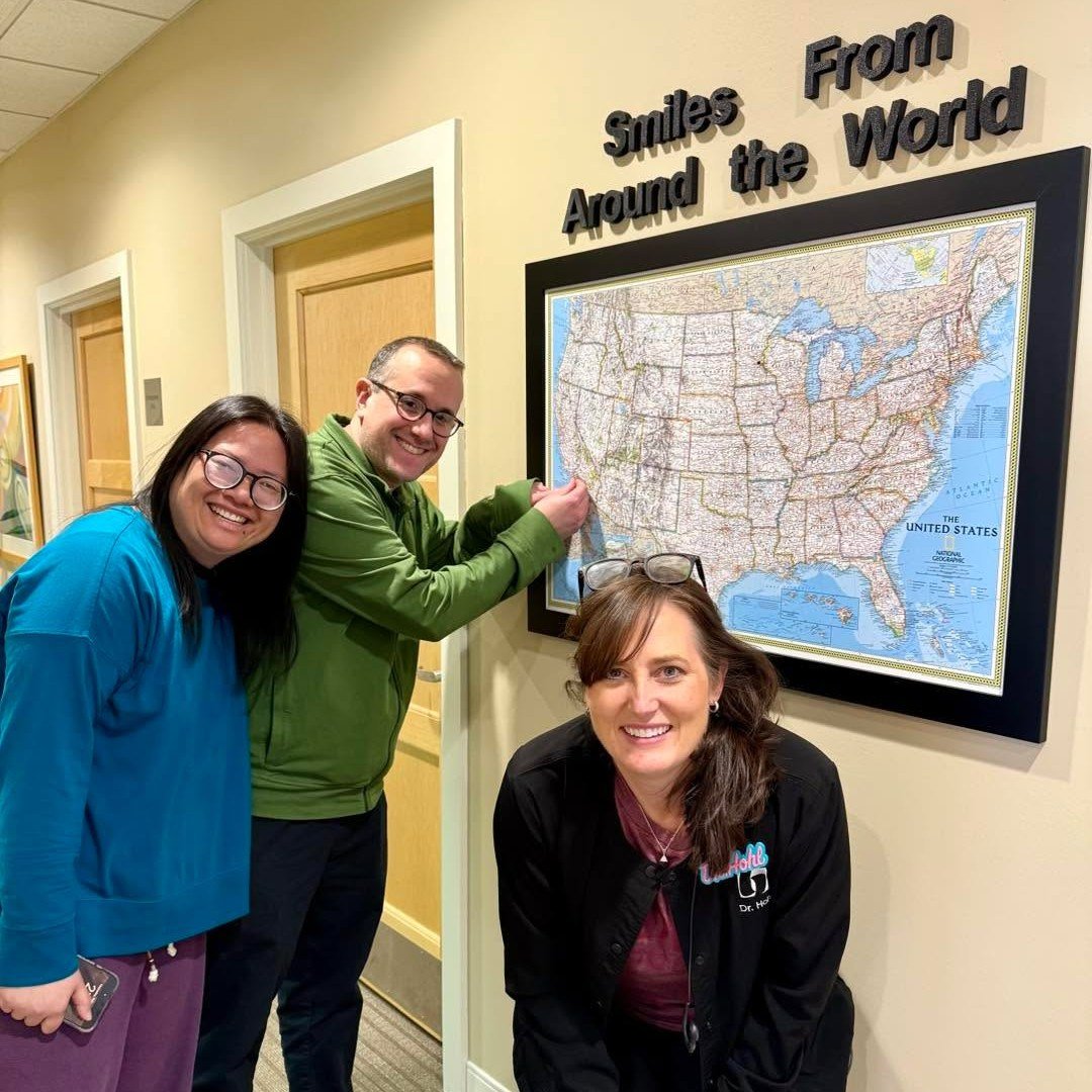 Three smiling people, two women and one man, are standing in front of a map of the United States on a wall. The man is pinning or pointing to the map while the women pose next to him. Above the map, there's a sign that says, 'Smiles Around the World.'