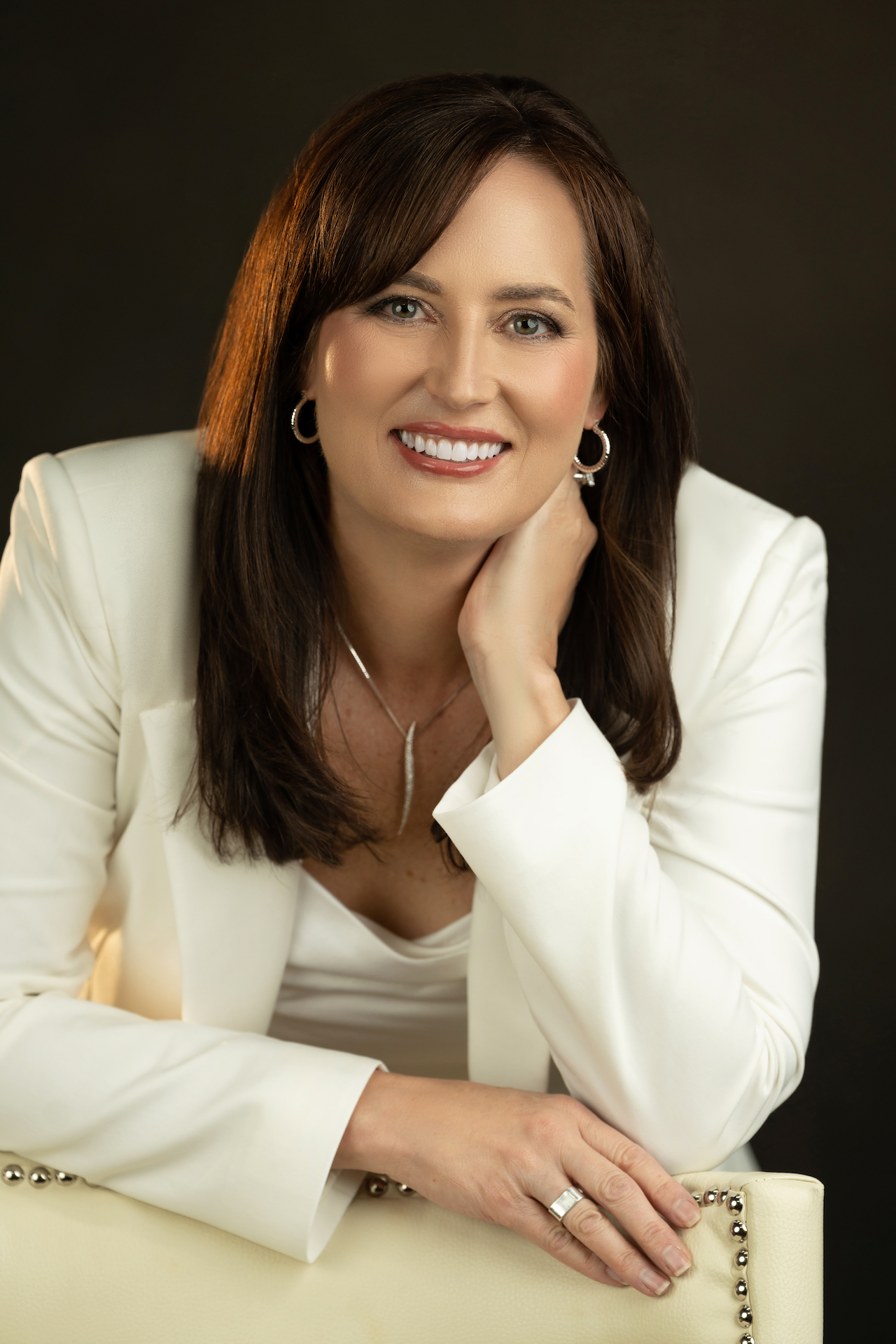 A woman with brown hair and blue eyes smiling, wearing a white blazer and jewelry, leaning on a cream-colored chair against a dark background.