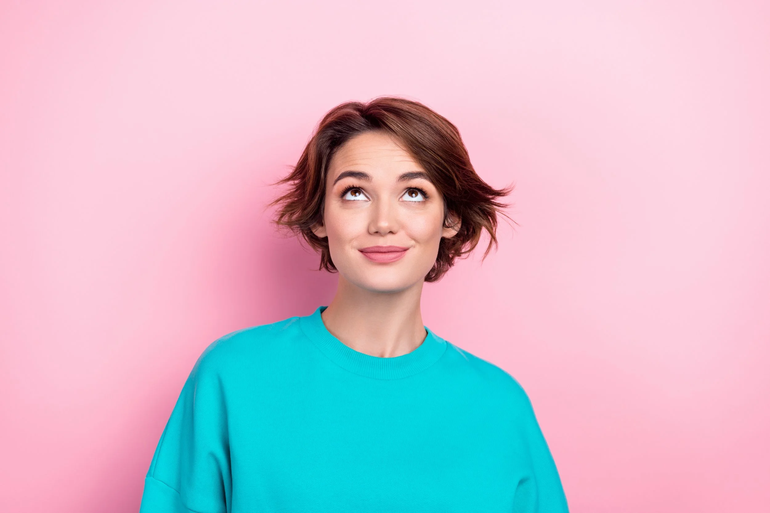 A woman with short, wavy brown hair wearing a bright blue shirt, smiling and looking upward, against a pink background.