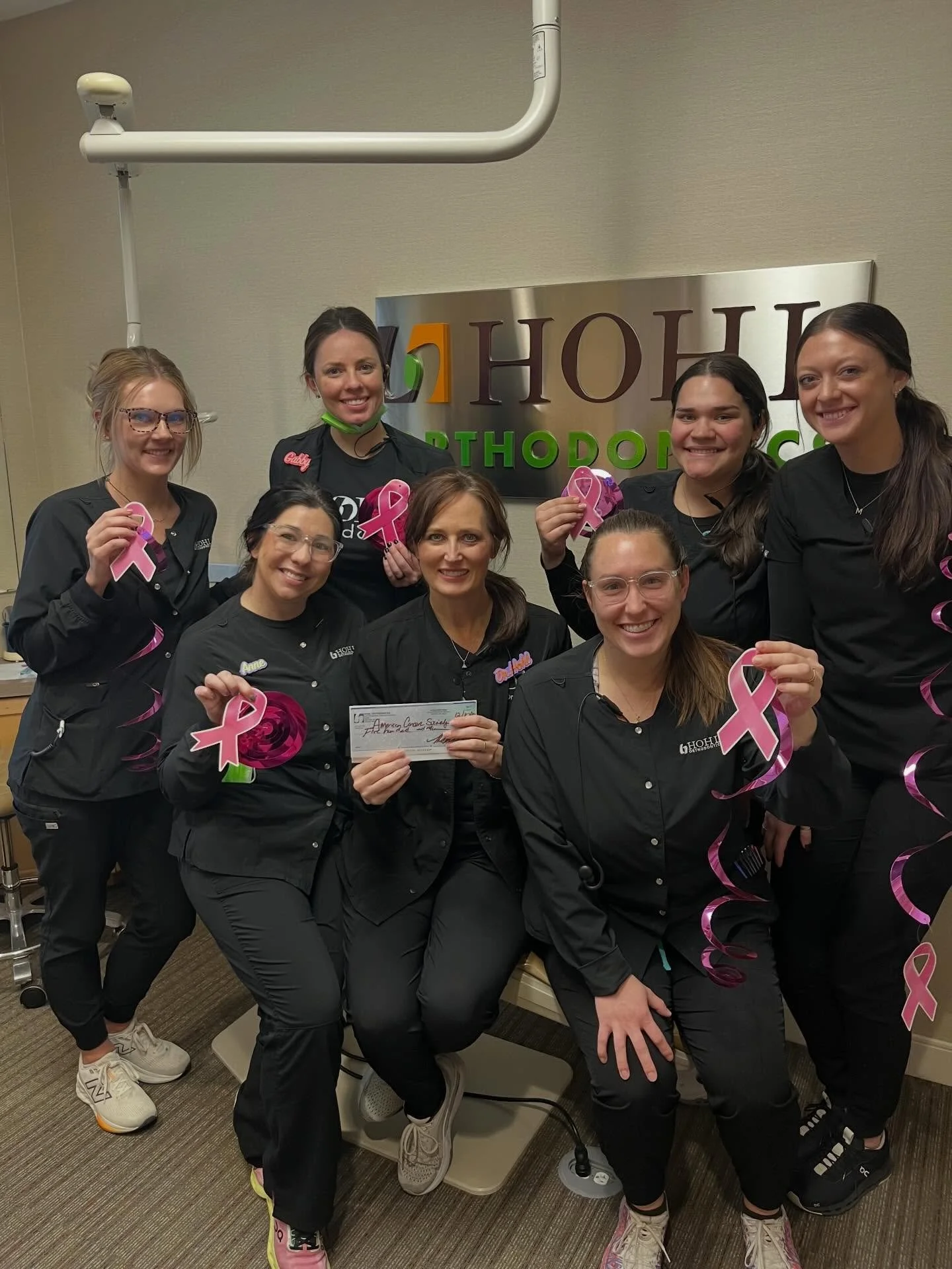 A group of eight women wearing black scrubs holding pink breast cancer awareness ribbons, standing in front of a sign that reads 'HOH Pediatric Dentistry,' inside a medical office. Some women are seated, and one is holding a check.