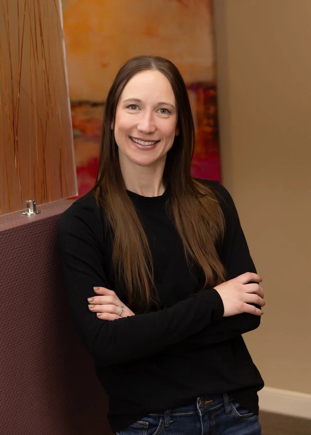 A woman with long brown hair, wearing a black long-sleeve shirt and jeans, smiling and standing with her arms crossed in a warmly decorated room.