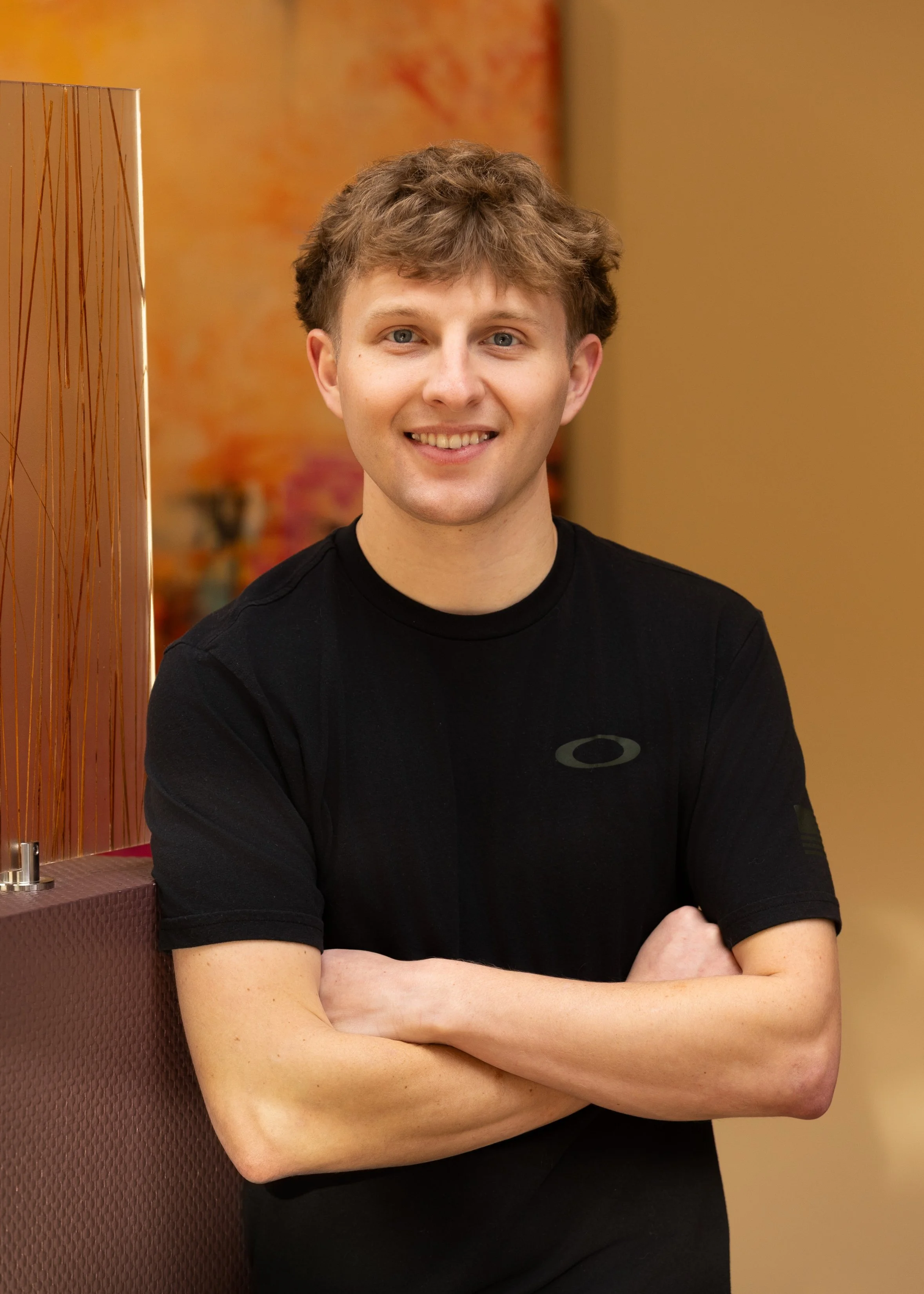 A young man with light brown, curly hair and blue eyes smiling, sitting with arms crossed, wearing a black t-shirt with a logo on the chest, indoors with warm lighting and abstract art on the wall in the background.