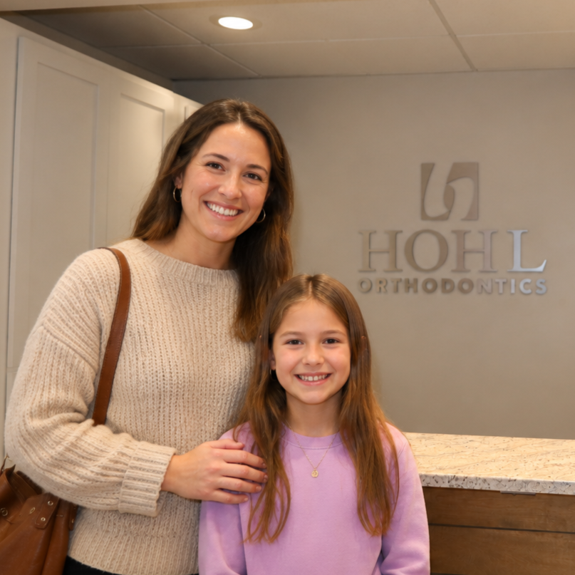 A smiling woman and girl standing in front of a wall with 'HOHL Orthodontics' logo, at a dental office reception area.