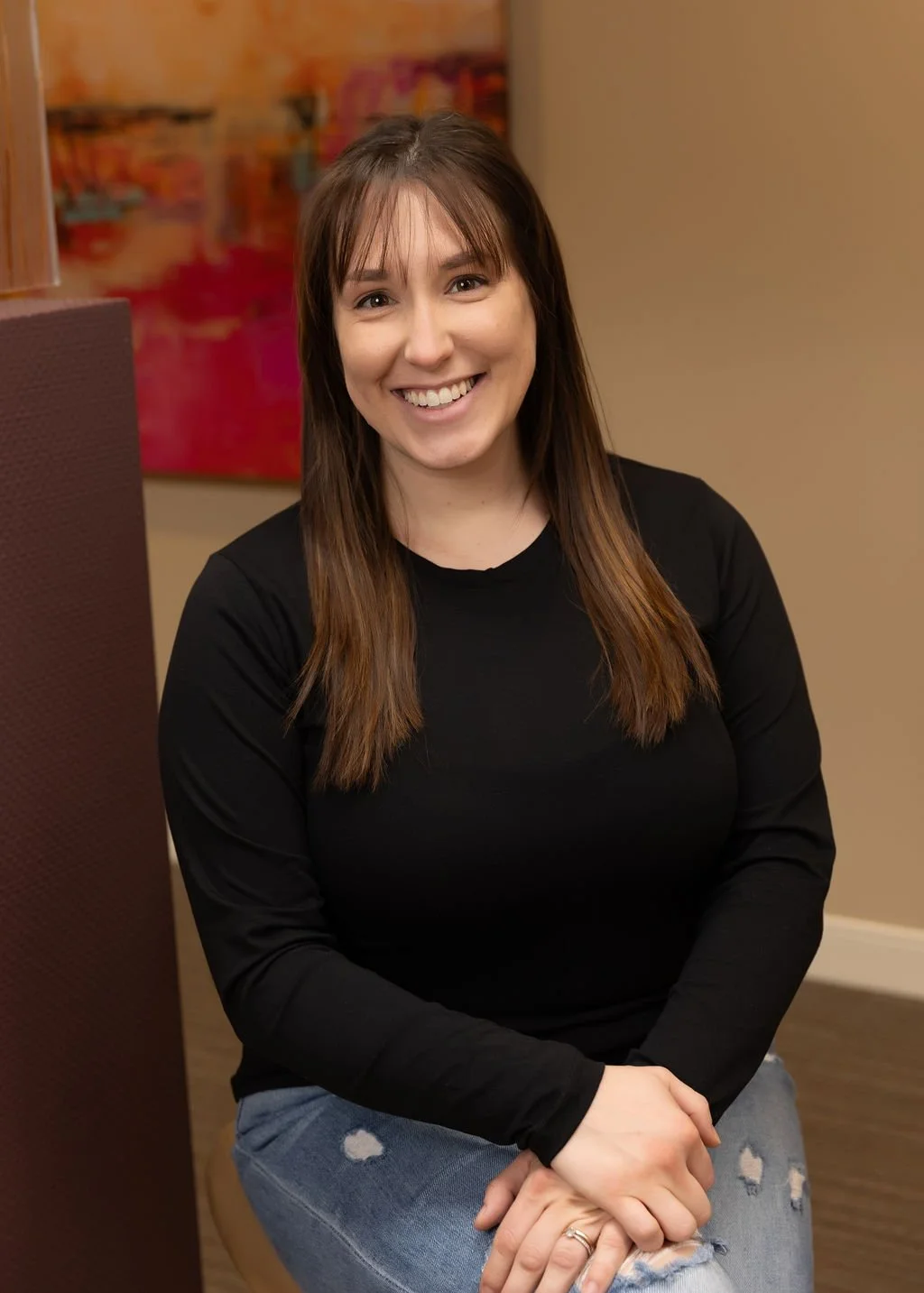 A smiling woman with long brown hair sitting on a chair, wearing a black long-sleeve shirt and ripped jeans, with colorful abstract art in the background.