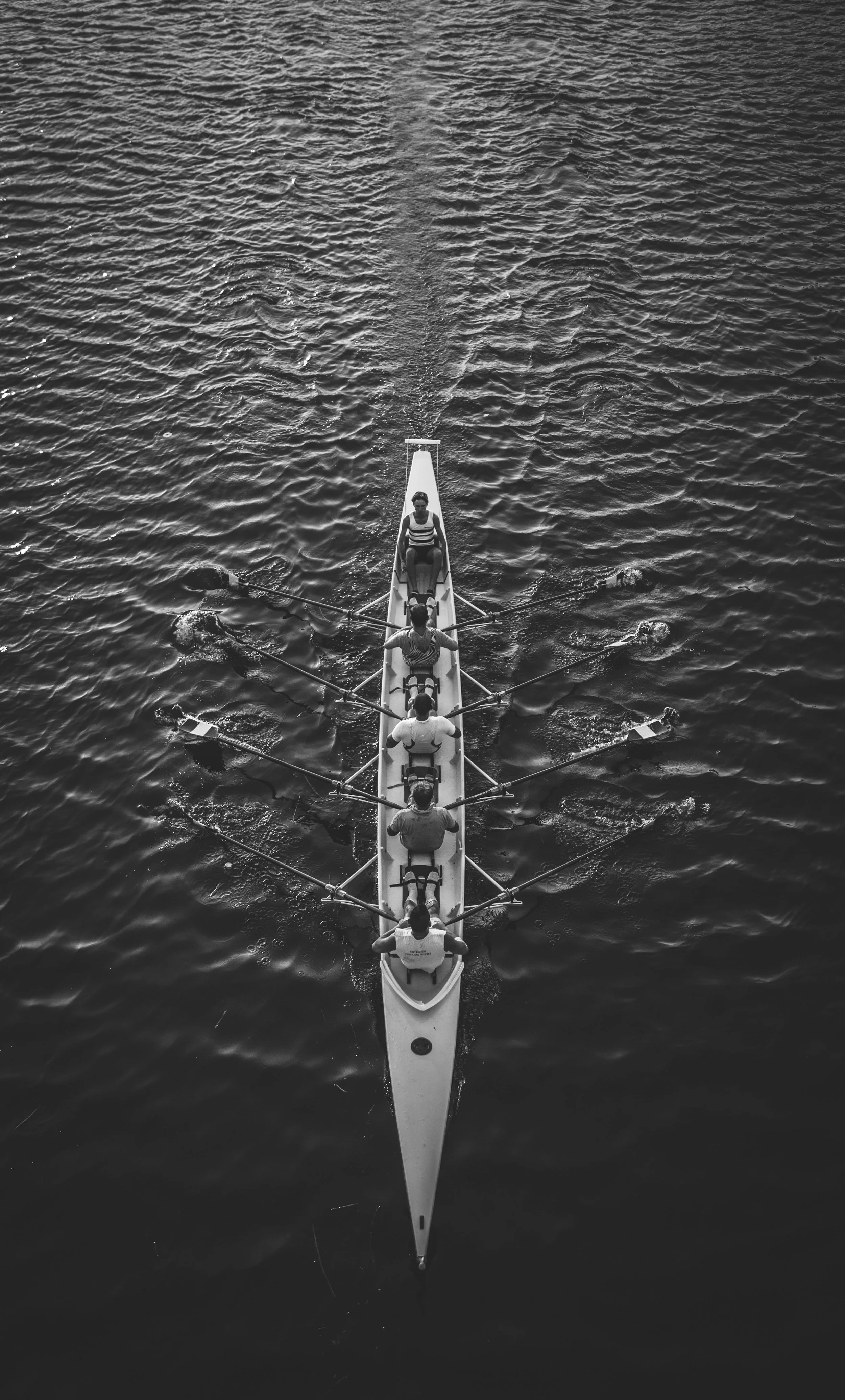 Black and white aerial view of five people rowing a long, narrow boat on calm water.