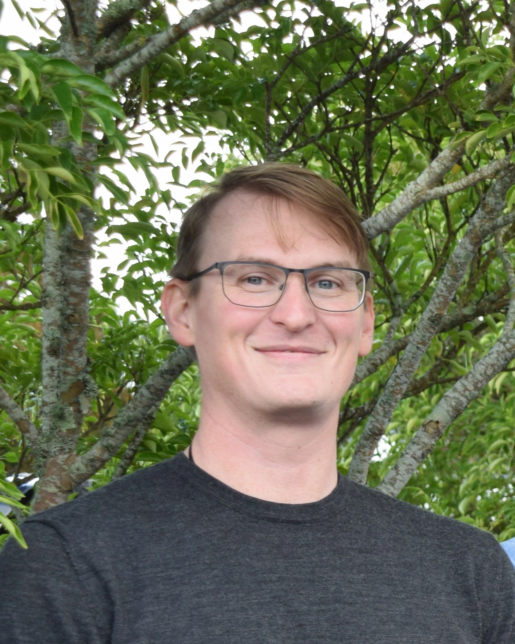 A young man with glasses and light brown hair, smiling, standing outdoors in front of a leafy tree.