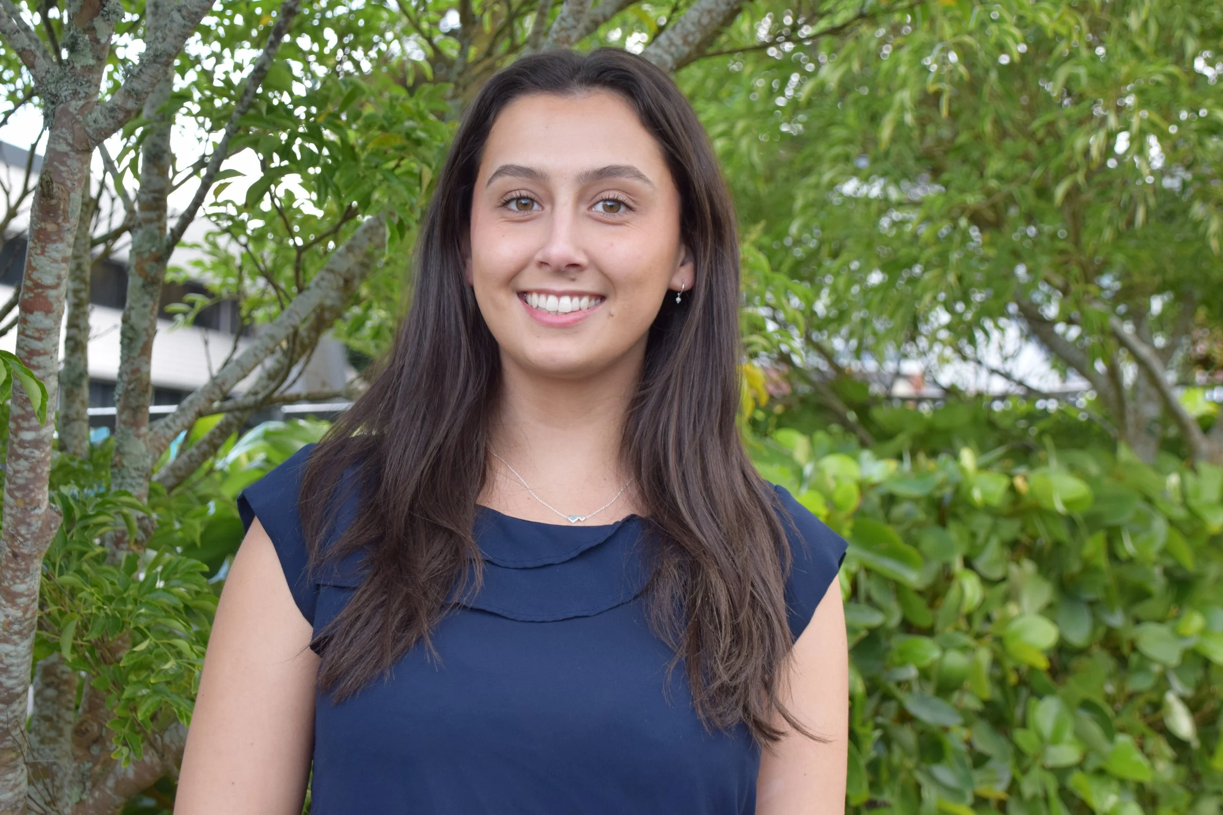 A young woman with long dark hair smiling outdoors, wearing a navy blue sleeveless top and small earrings, with green trees and shrubs in the background.
