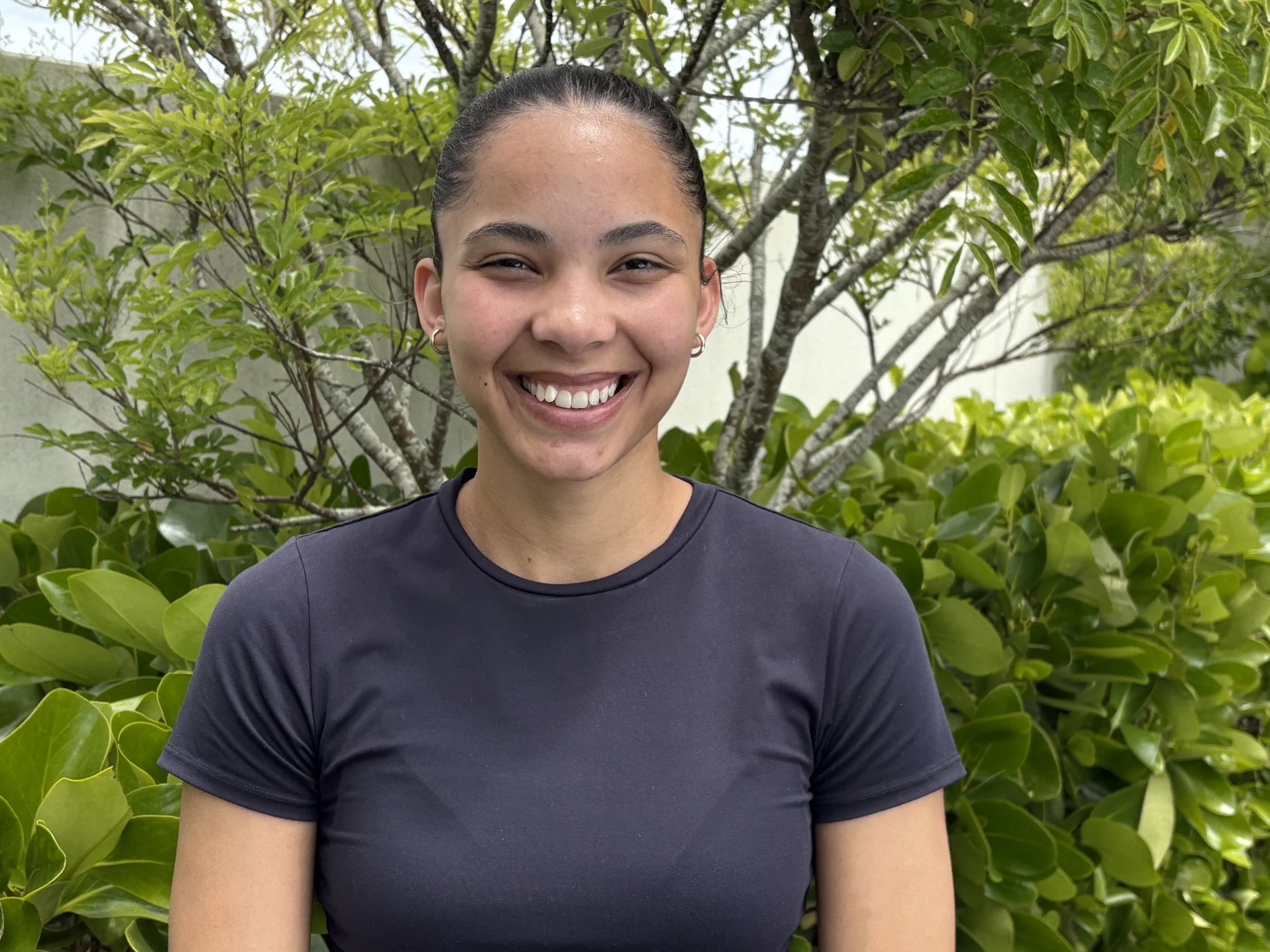 A young woman with dark hair pulled back, smiling, standing outdoors in front of green shrubbery and trees.