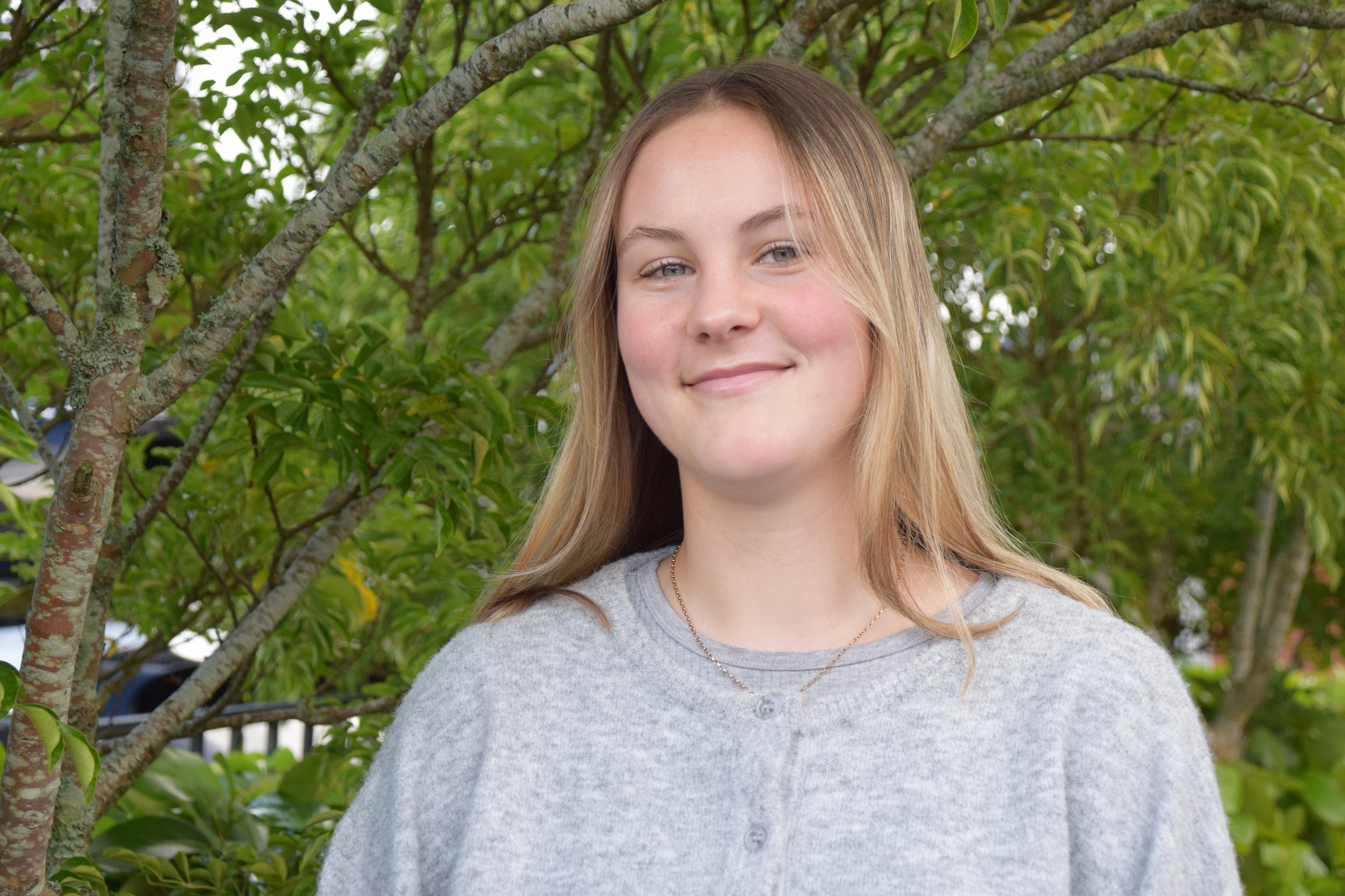 A young woman with long blond hair smiling outdoors with green trees in the background.