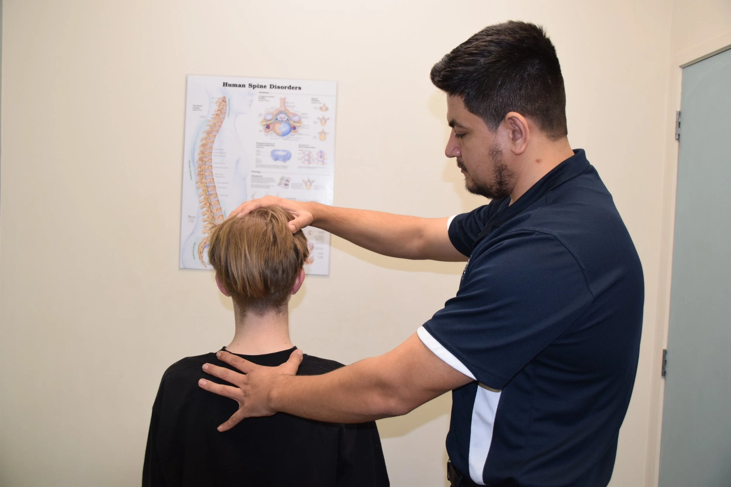 A male healthcare professional examining a woman's neck and head in a medical office, with a chart on human spine disorders on the wall.
