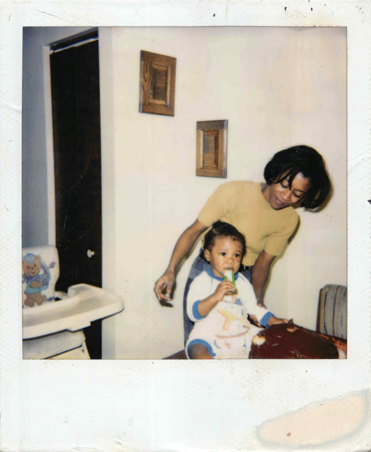 A woman and a young boy in pajamas in a room. The woman is leaning over the boy who is sitting on a table, holding a popsicle. There are framed pictures on the wall and a high chair to the side.