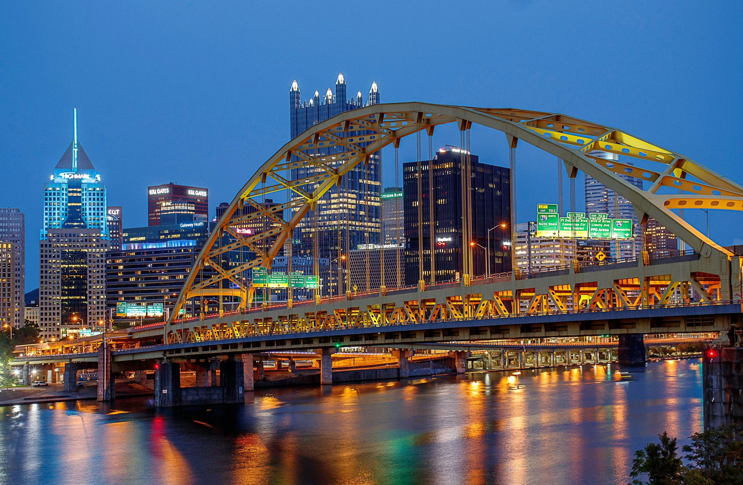 Night view of downtown Pittsburgh skyline with the yellow Roberto Clemente Bridge over the water in the foreground.