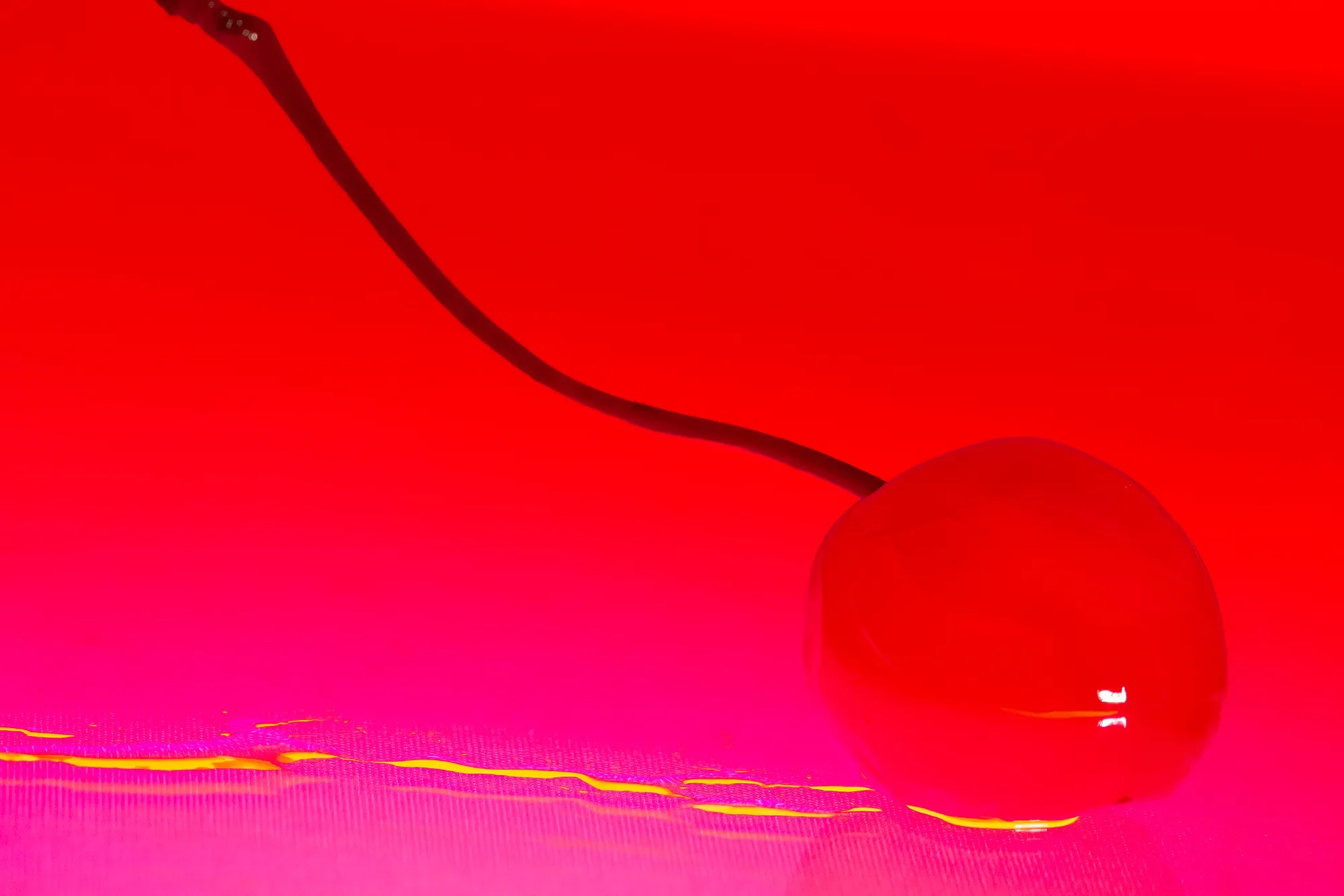 Close-up of a bright red cherry with a dark stem, resting on a pink and red surface with some yellow highlights.