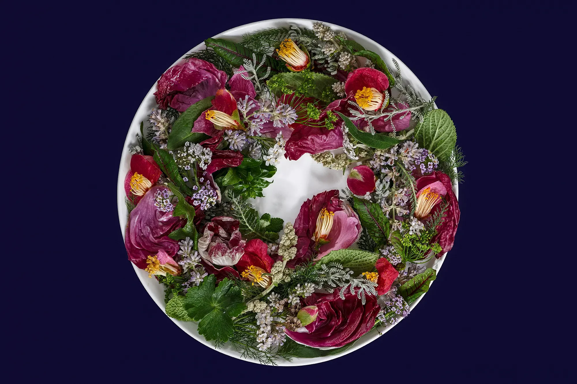 A round floral arrangement with pink, purple, and white flowers, including roses and small blossoms, on a white dish against a dark background.