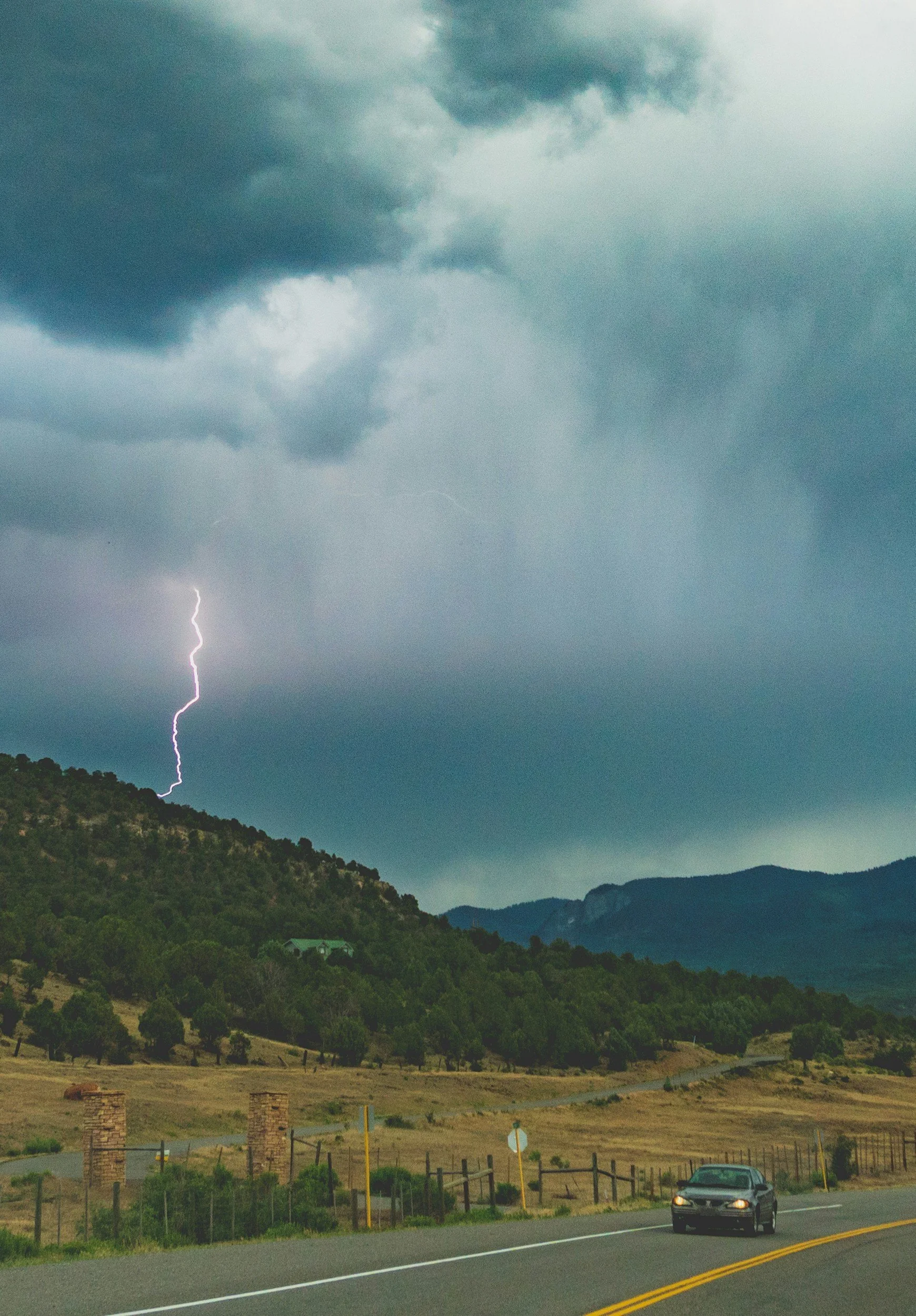 A car traveling on a mountain road during a thunderstorm with lightning striking in the distance and dark storm clouds overhead.