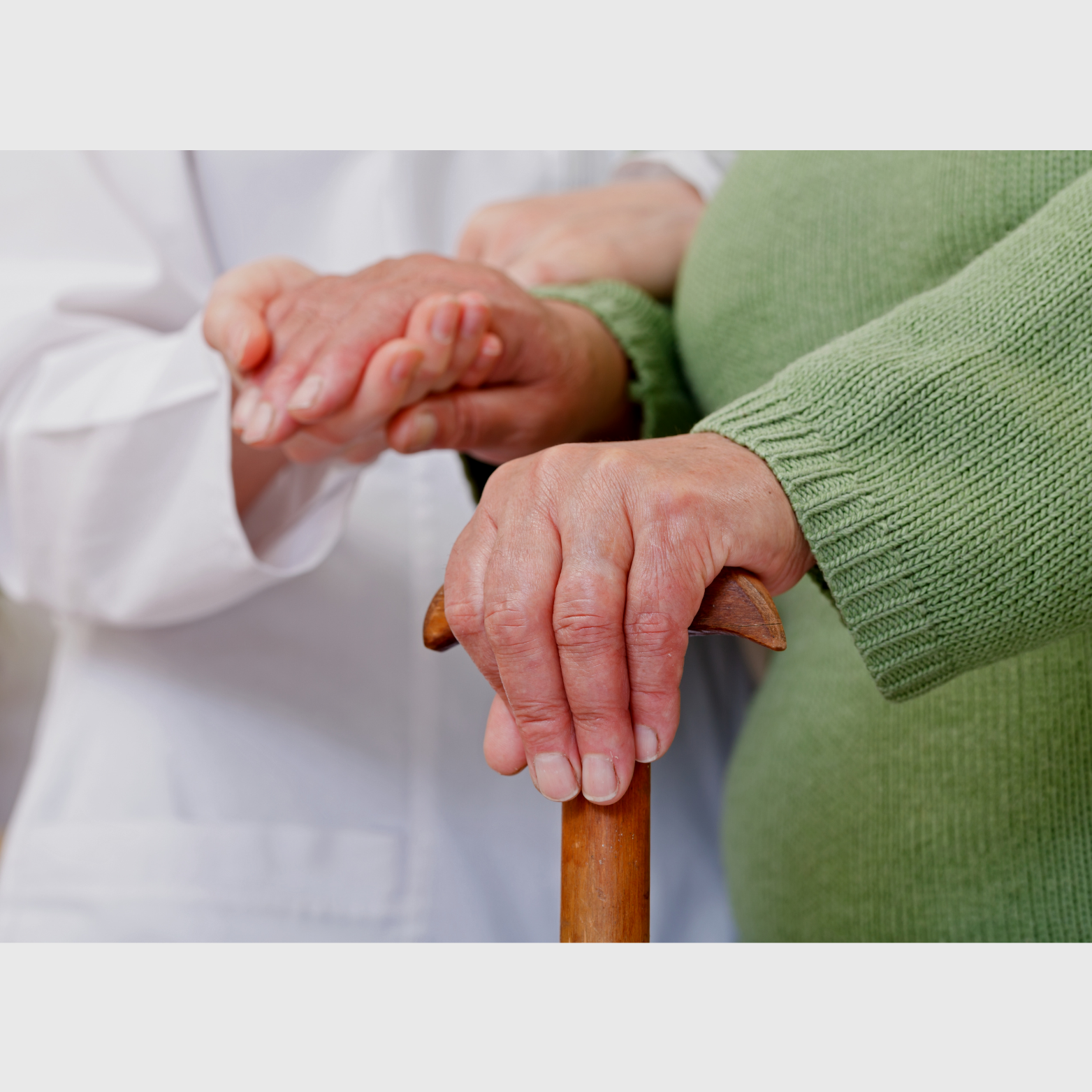 A healthcare professional holding an elderly woman's arm in a supportive manner with one hand, while the elderly woman rests her hand on a wooden cane, wearing a green sweater.