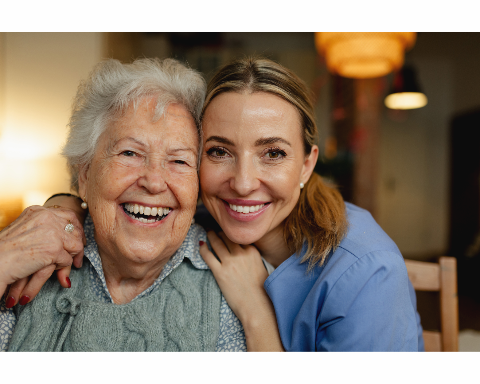 A young female healthcare professional and an elderly woman smiling and slightly hugging at home.